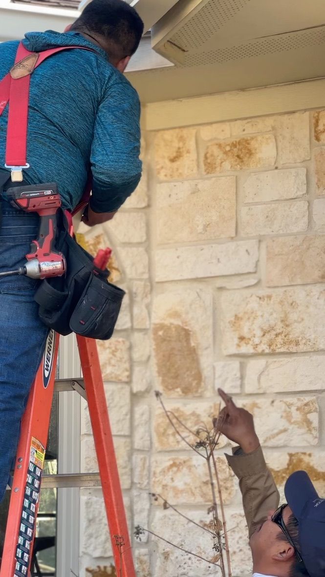 Two workers, one on a ladder, point at a stone wall. One wears a blue shirt and red suspenders.