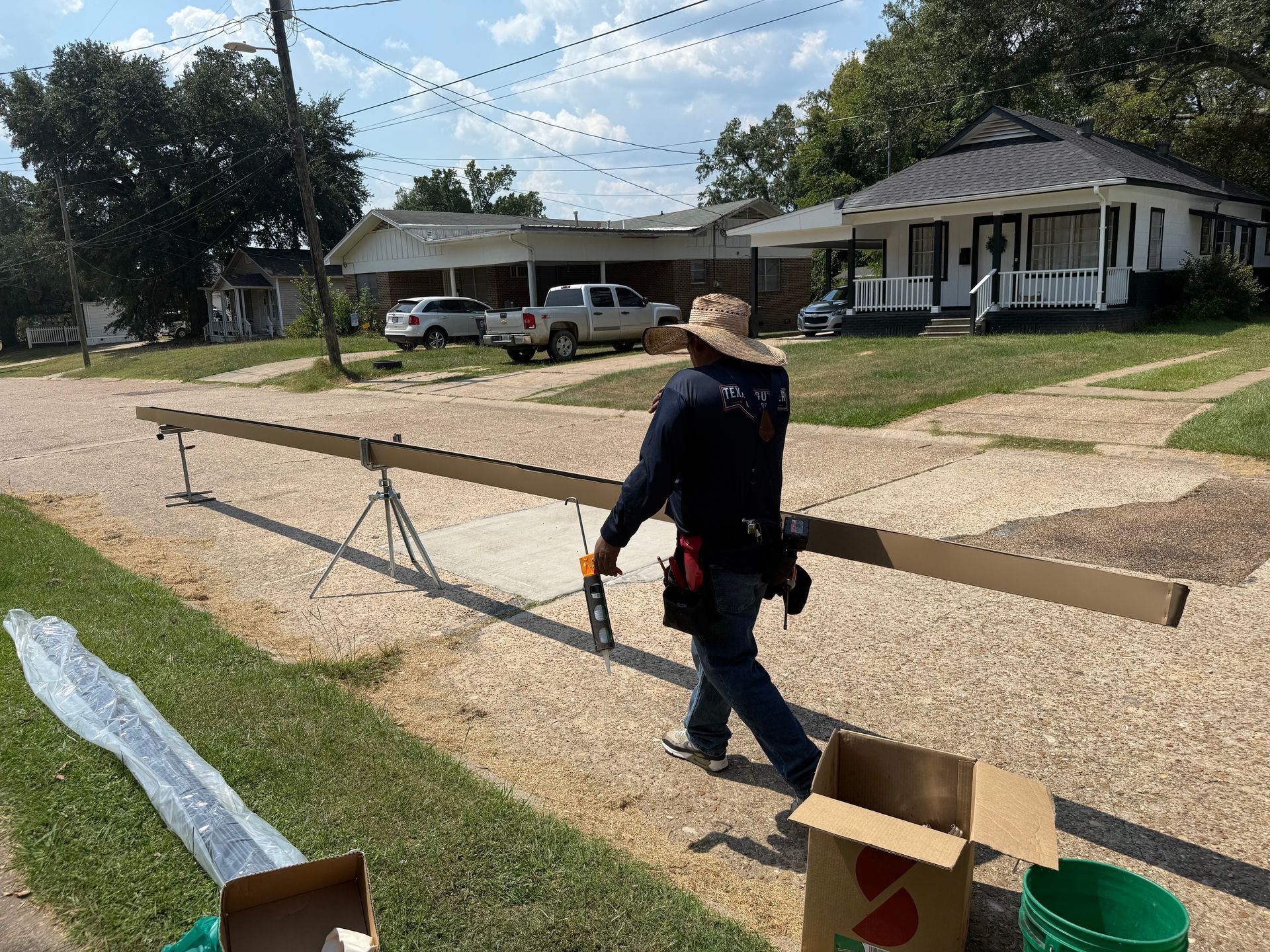 Person carrying long wooden beams, setting them on a gravel driveway. Houses and sky in background.