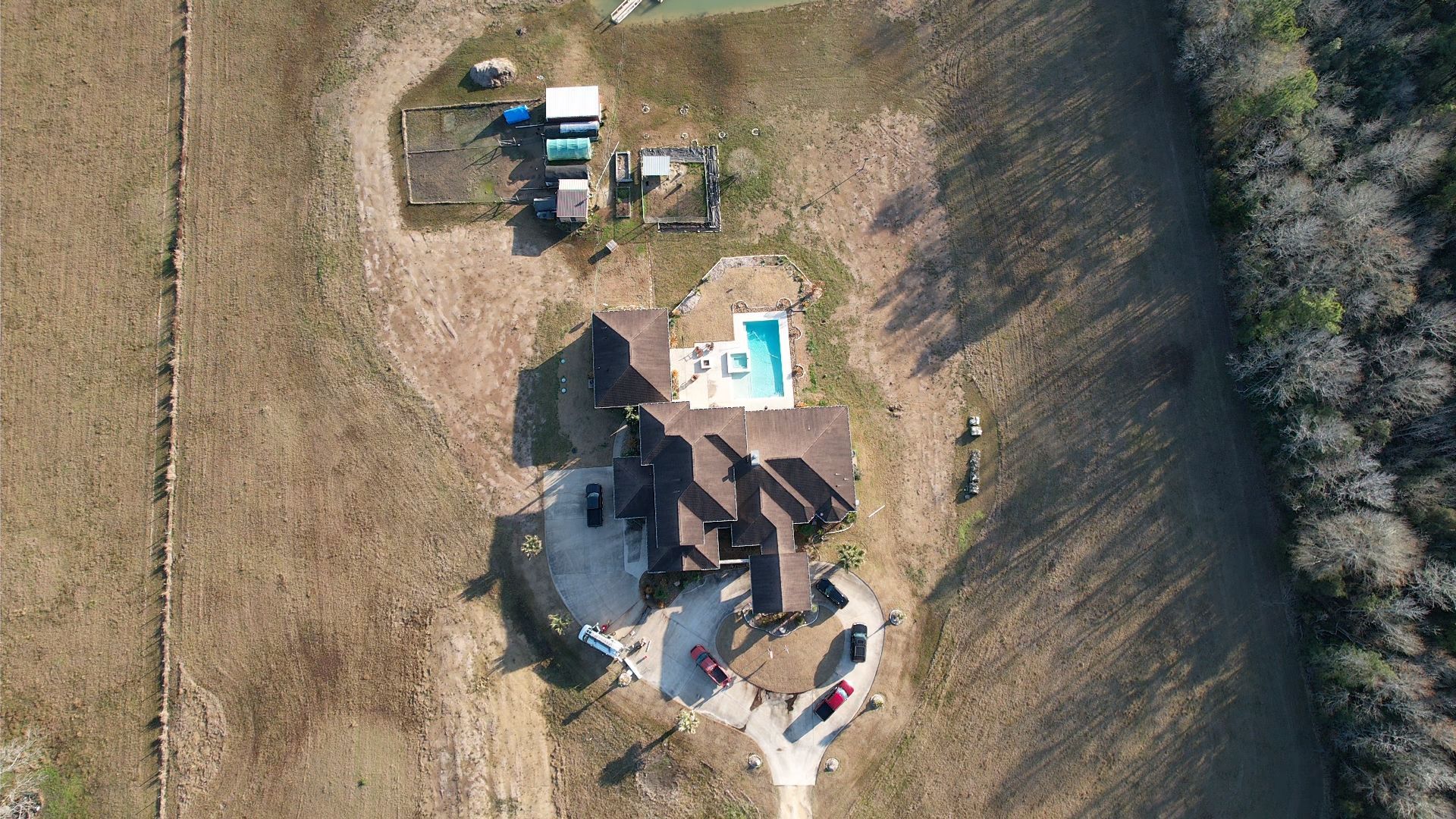 Aerial view of a house with a pool, vehicles, and surrounding brown field and trees.