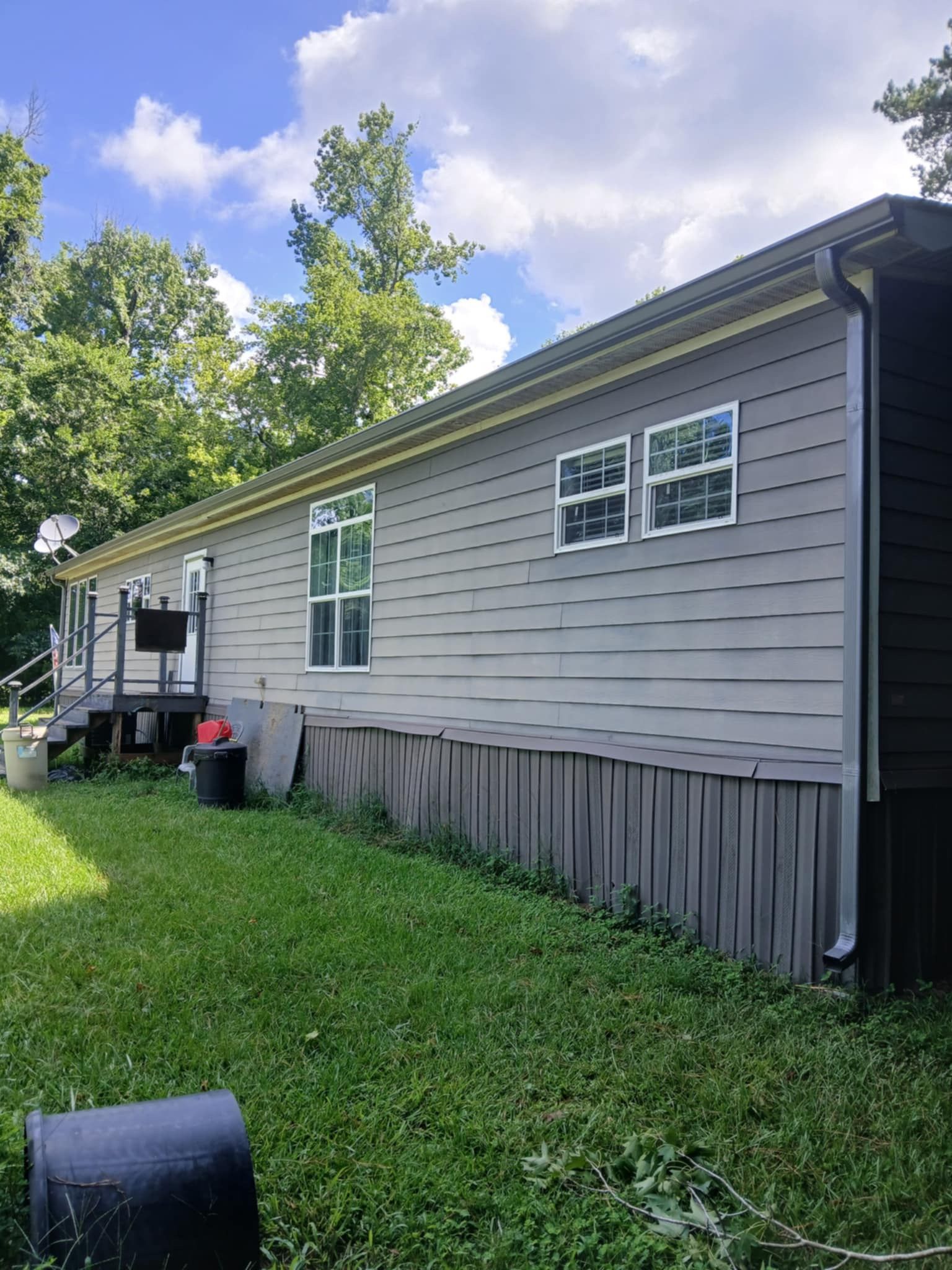 Side view of a light gray house with two white-framed windows, surrounded by green grass and trees.