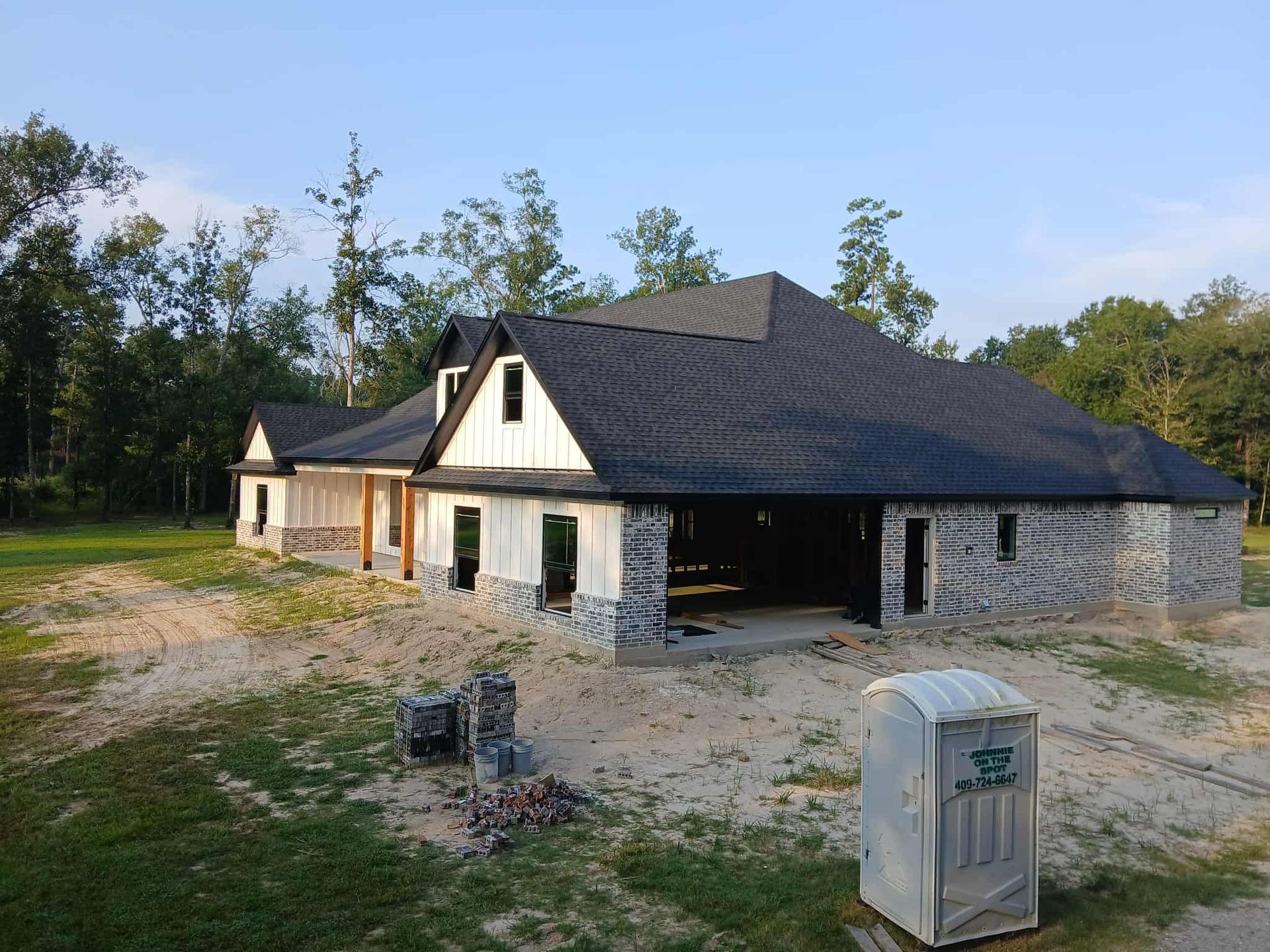 House under construction with a dark shingle roof and stone facade. Porta-potty in front.