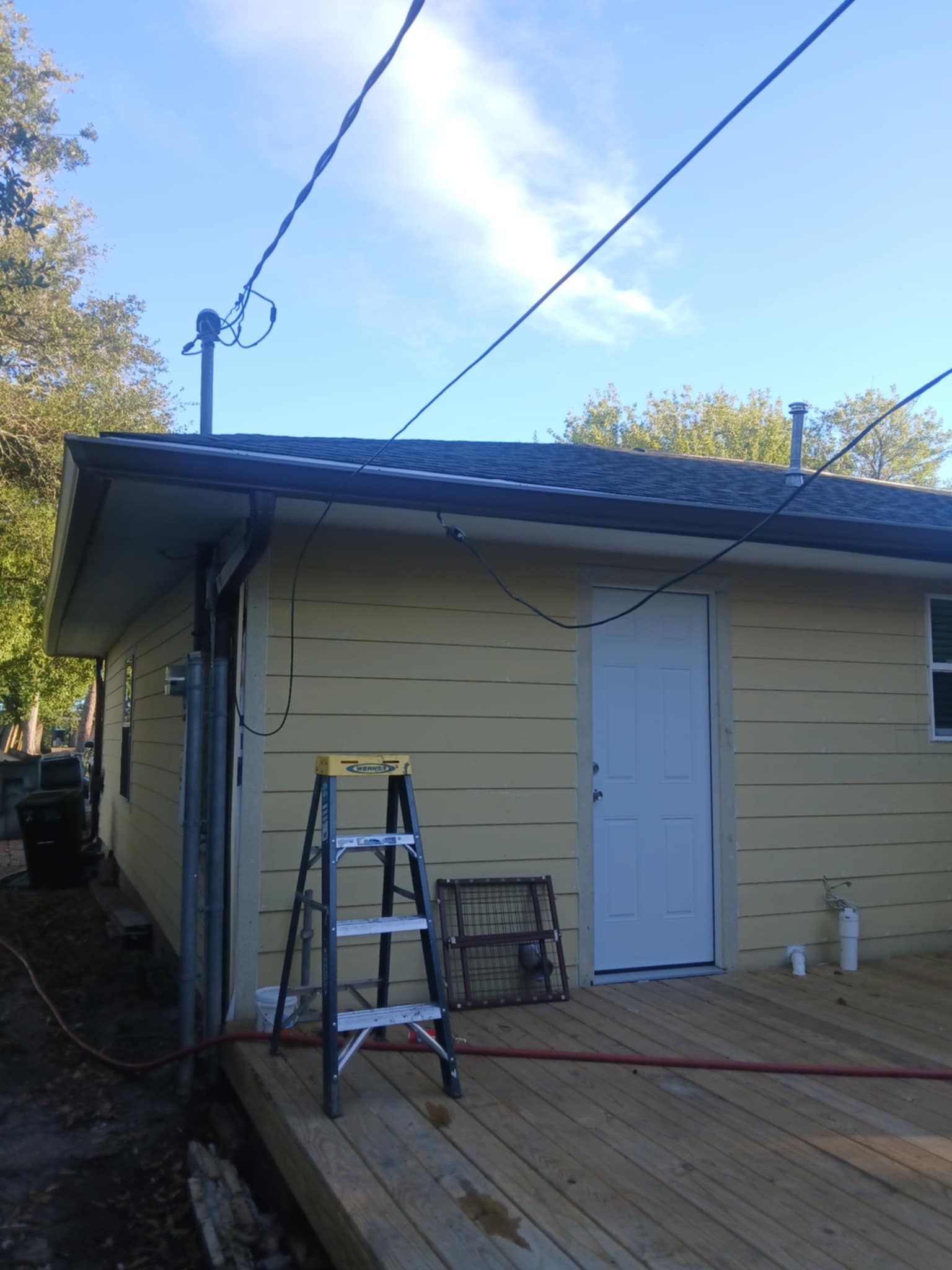 Yellow house with white door, wooden deck, ladder, and power lines against a blue sky.