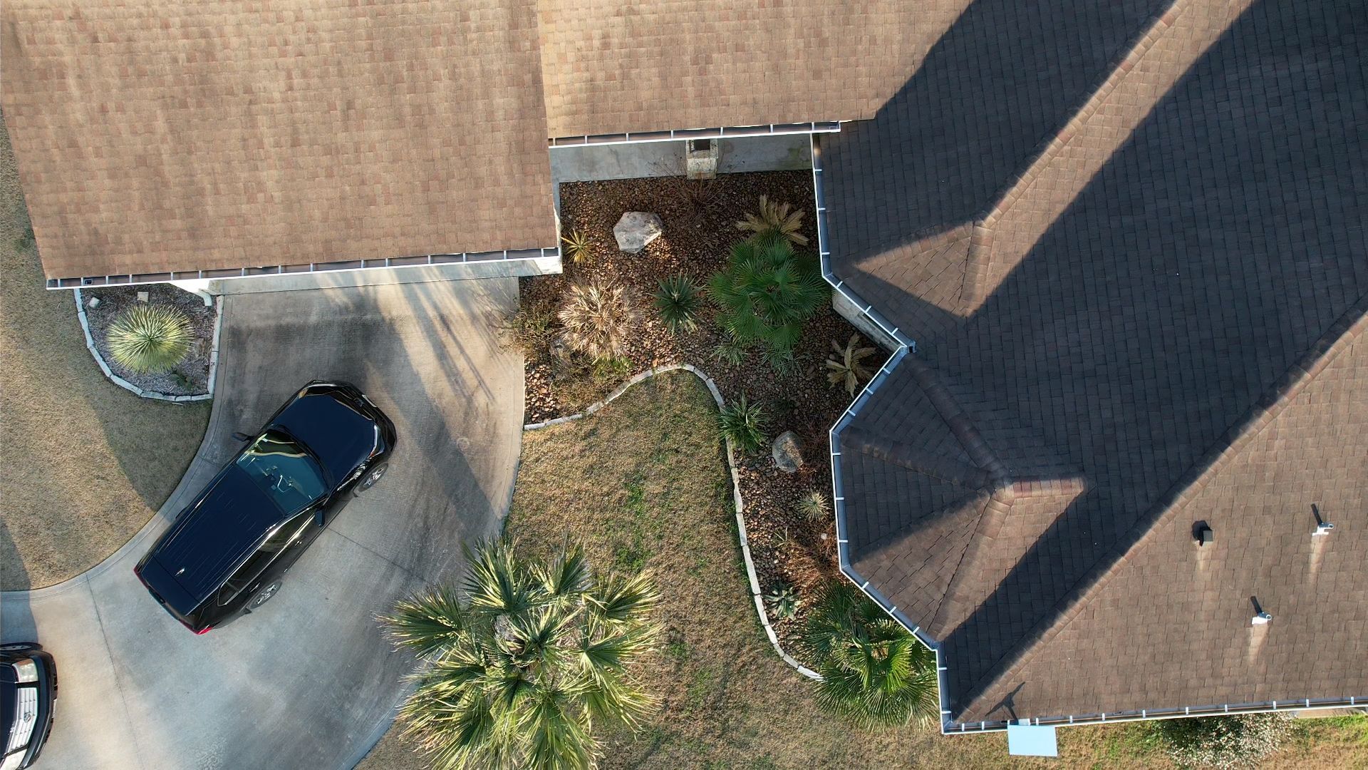 Overhead view of a house with a driveway and car, brown roof, and garden area with plants.