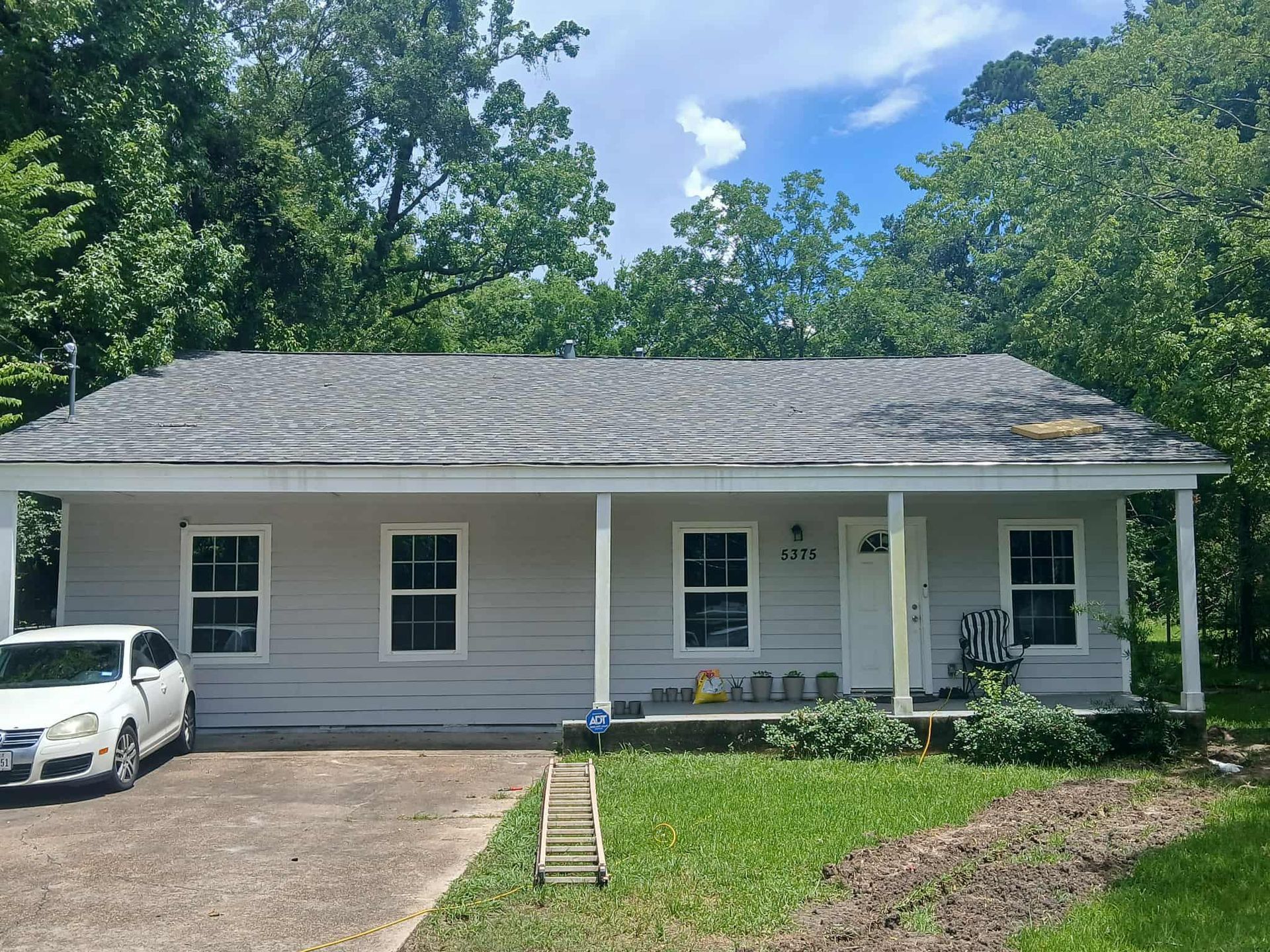 Small, light-colored house with worn roof and porch. A car is parked in the driveway. Trees and sky in the background.