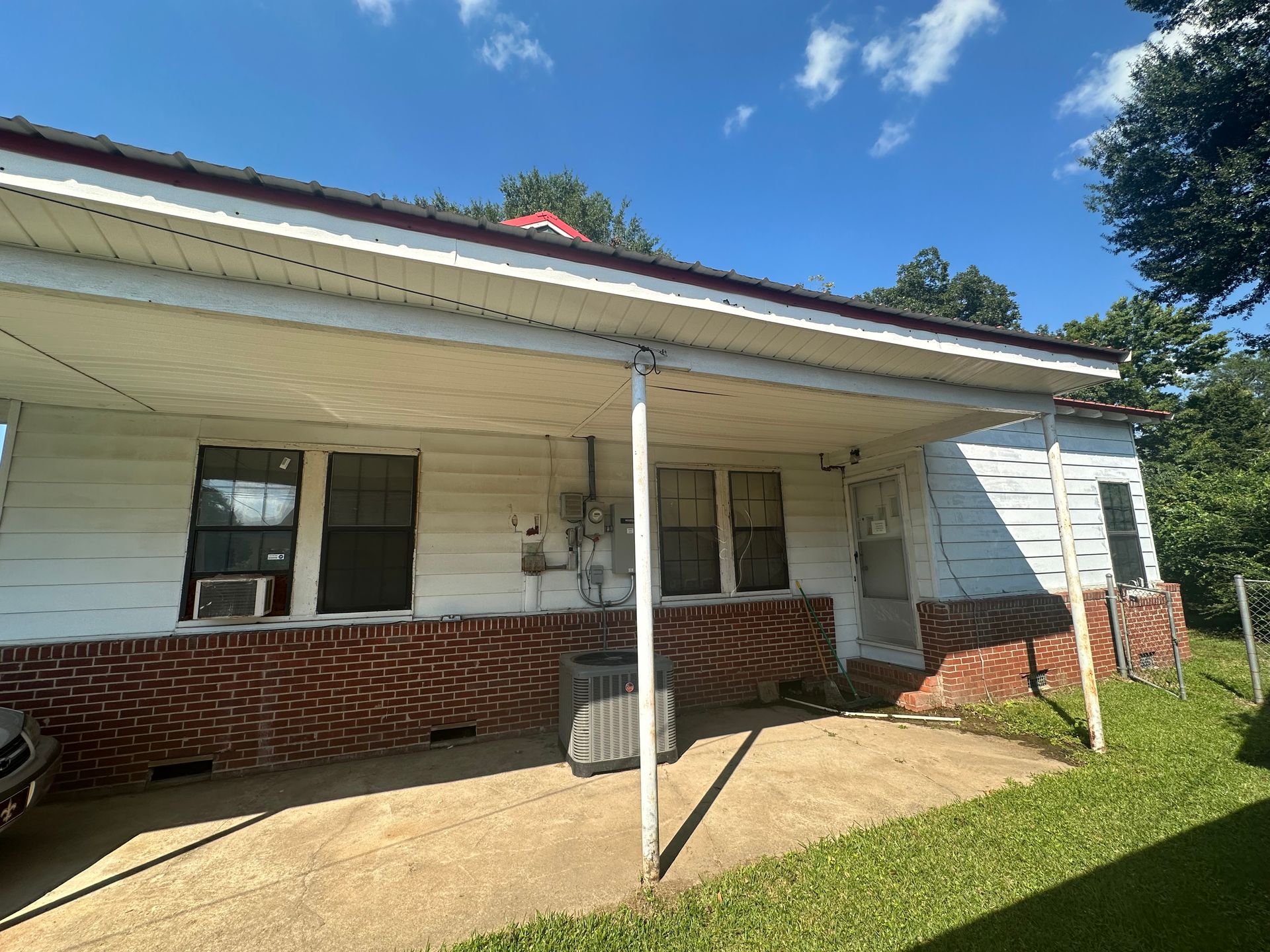 White house with red brick foundation, carport, and green lawn under a blue sky.