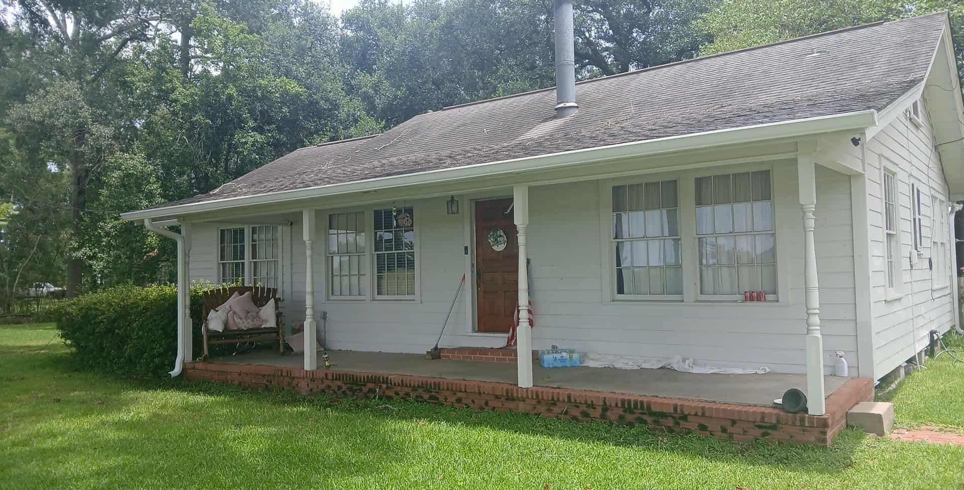 White, one-story house with porch, brick foundation, green grass, and trees. Brown door, windows, and chimney.