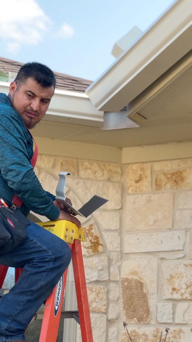 Man on ladder installing a security camera under a roof's overhang, using tools. Exterior with stone wall and blue sky.