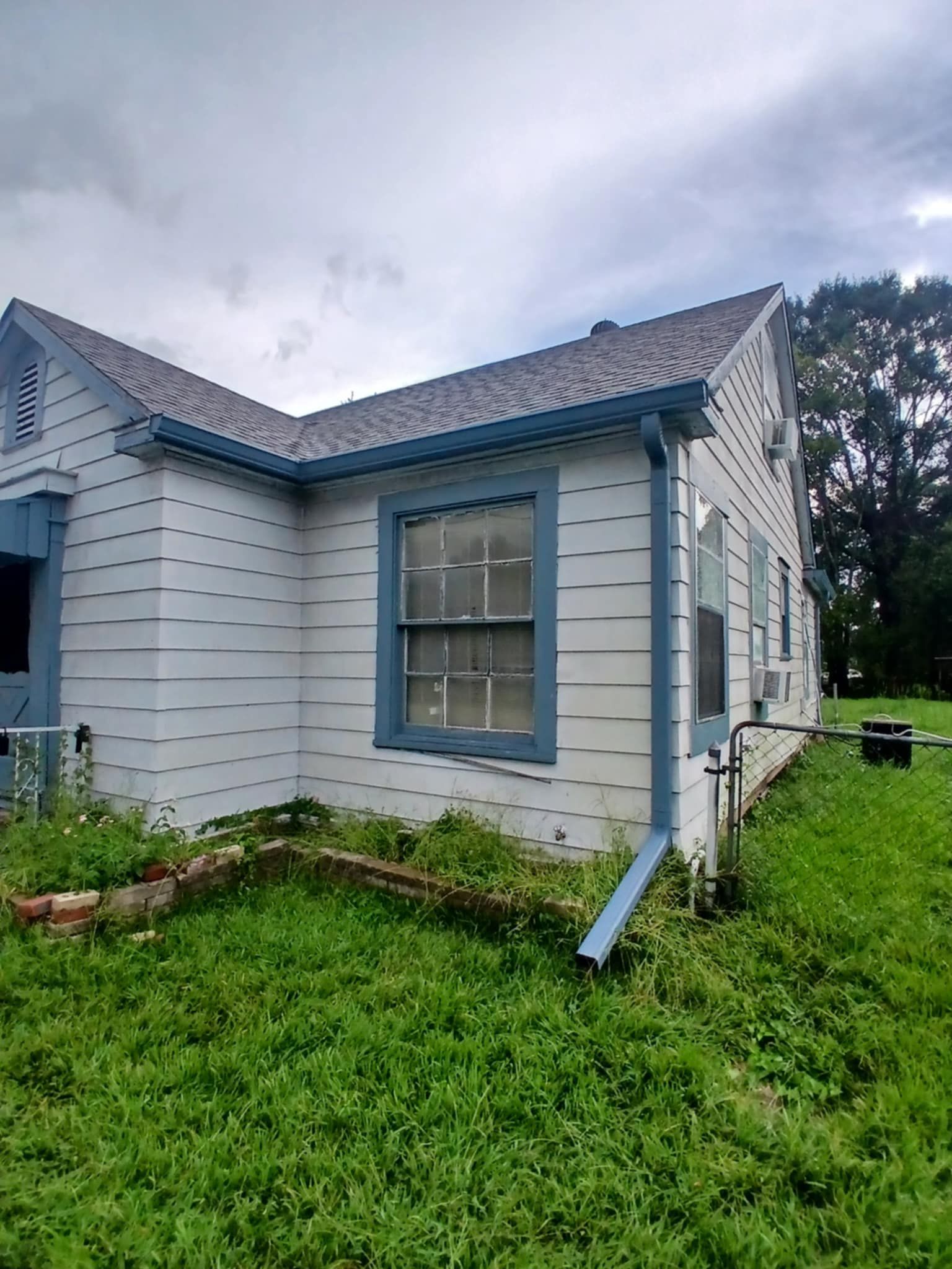 White house with blue trim, overgrown grass, cloudy sky.