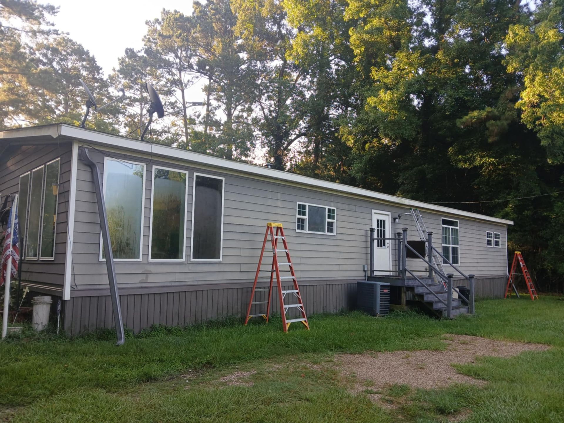 Gray mobile home with tall windows, two ladders, and a black staircase in a wooded area.