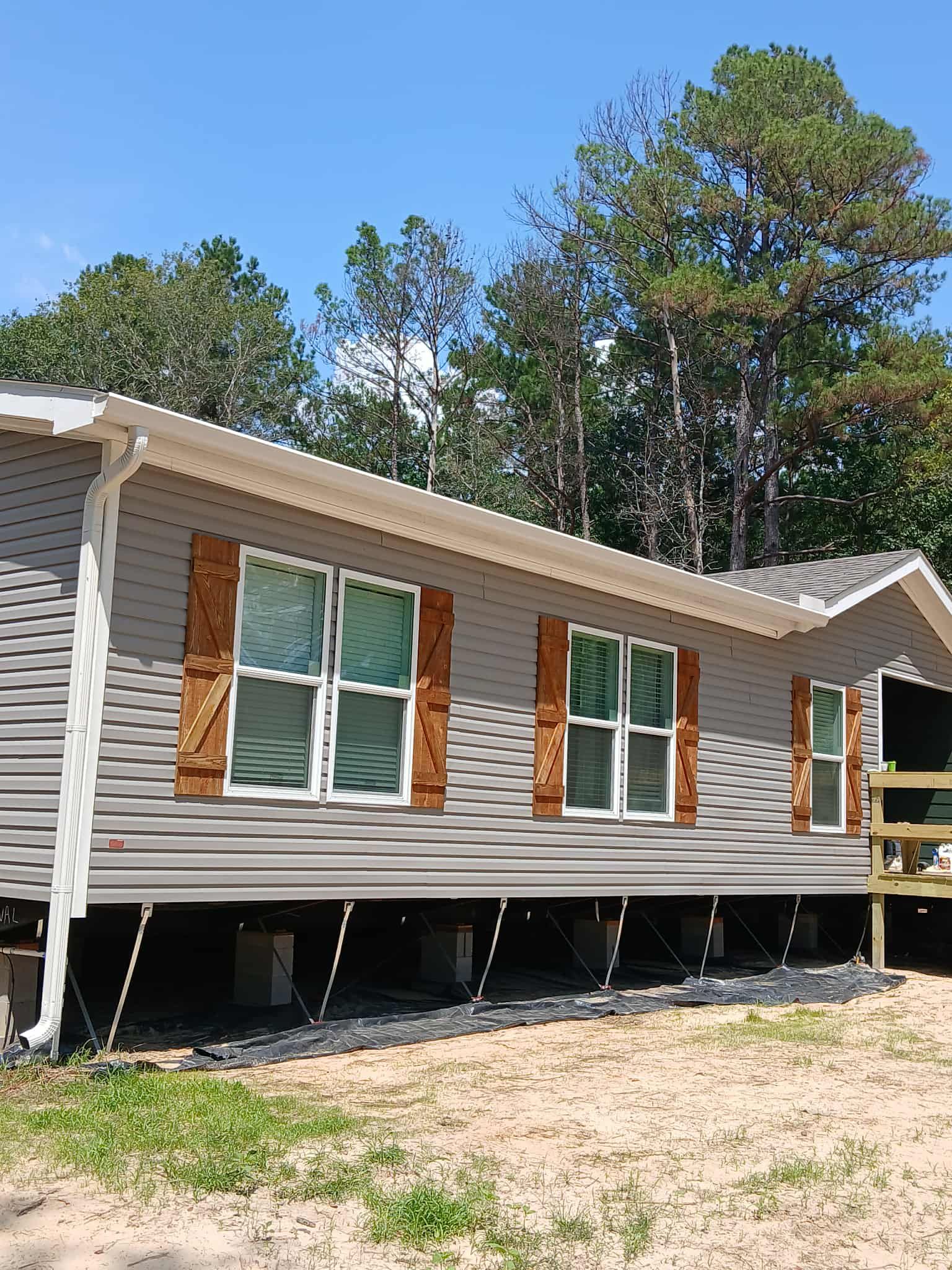 Gray house with brown shutters, raised on blocks, surrounded by trees and grass.