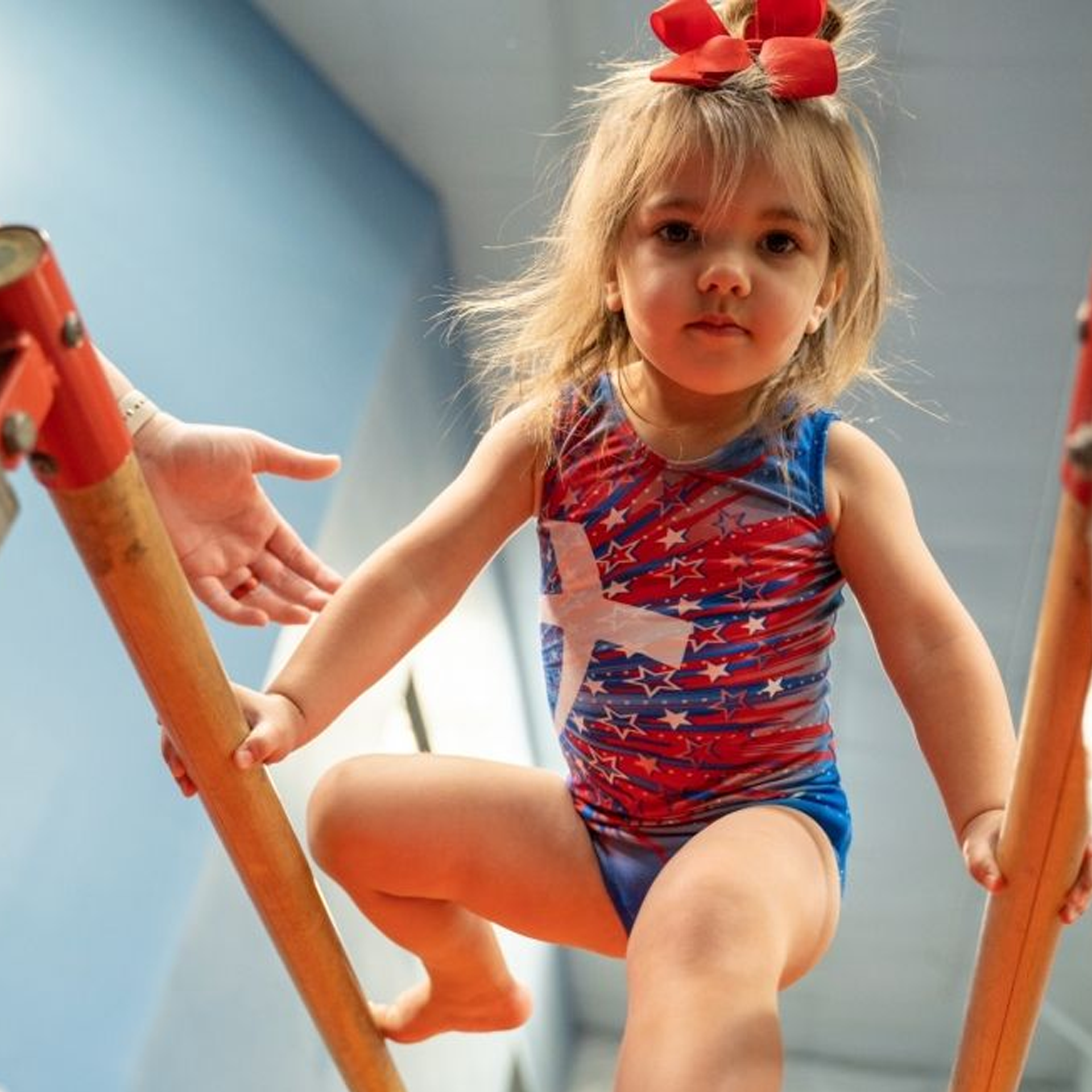 A little girl in a red white and blue leotard is sitting on a bar