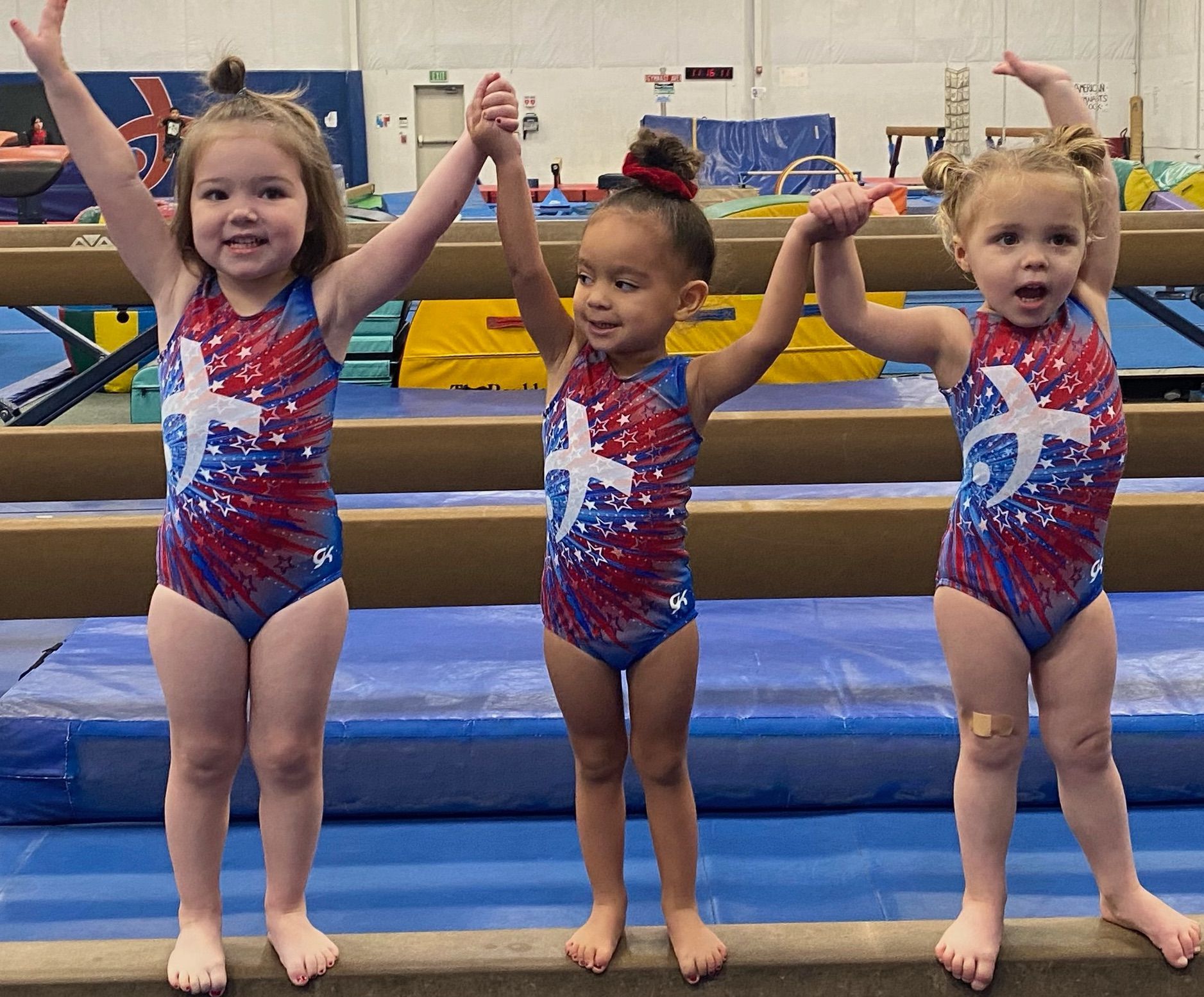 Three little girls are standing on a balance beam with their arms in the air.