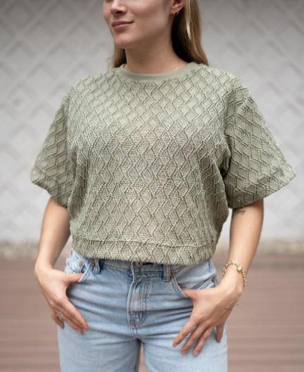 Una mujer que lleva una blusa de encaje verde y jeans azules está de pie con las manos en los bolsillos.