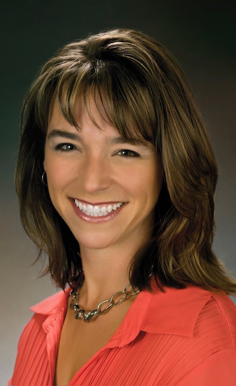 Woman with brown hair smiling, wearing an orange shirt and gold chain necklace.