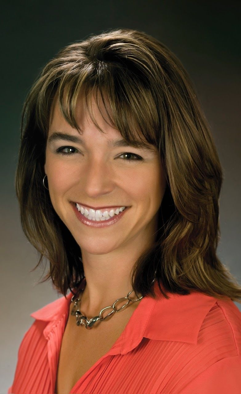 Woman with brown hair smiling, wearing an orange shirt and gold chain necklace.