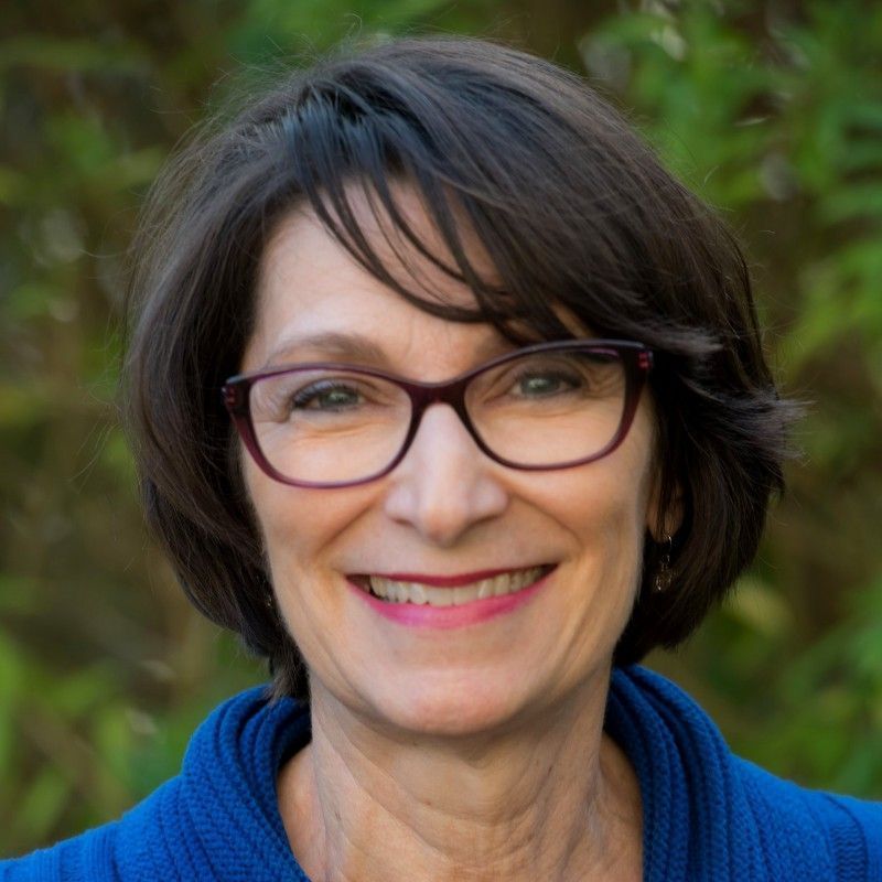 Woman with short dark hair, glasses, smiling, wearing a blue sweater, in a green outdoor setting.