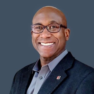 Man in glasses smiles, wearing a suit jacket and collared shirt against a gray background.