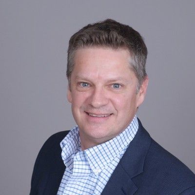 Man in blue blazer and patterned shirt smiling, headshot against gray background.