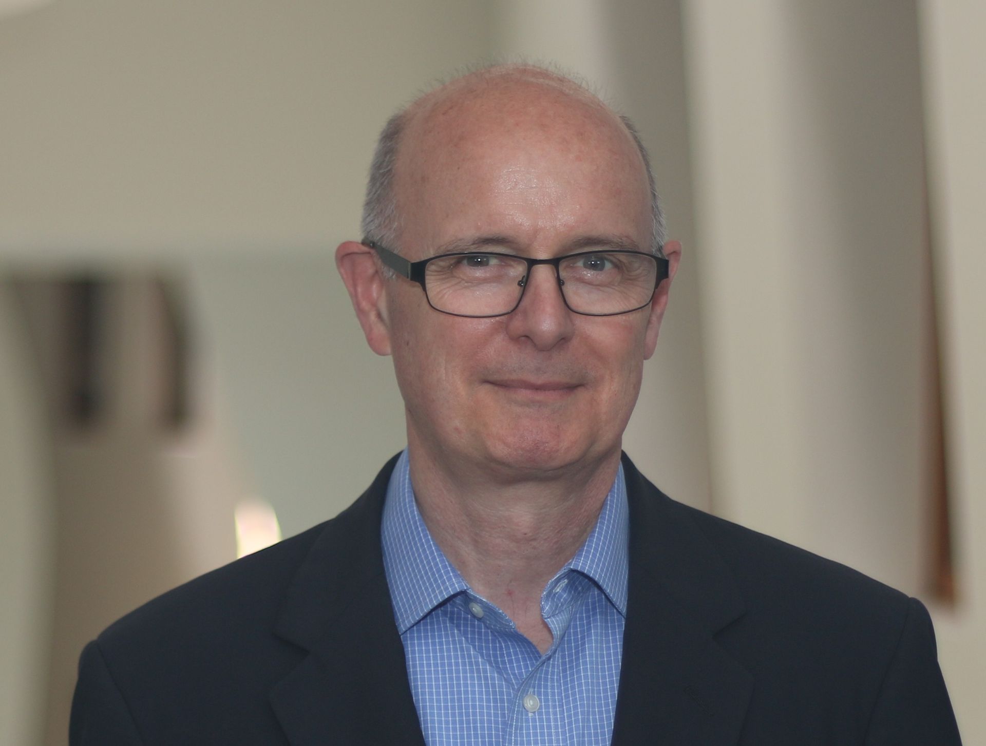 Man with glasses and blue shirt smiles, wearing a dark jacket, against a neutral-toned background.