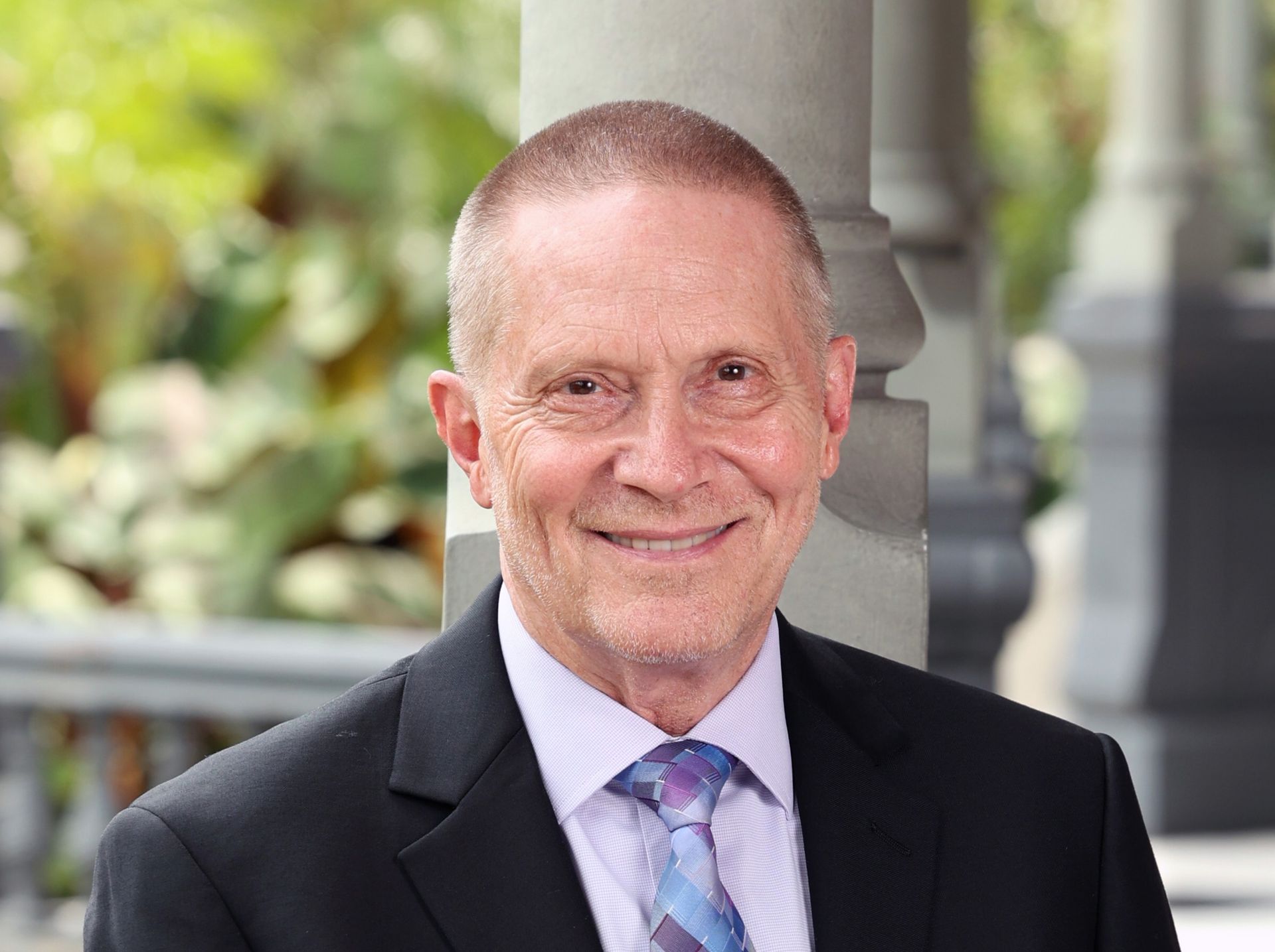 Man in a suit smiles, outdoors near a decorative pillar, with green foliage in the background.