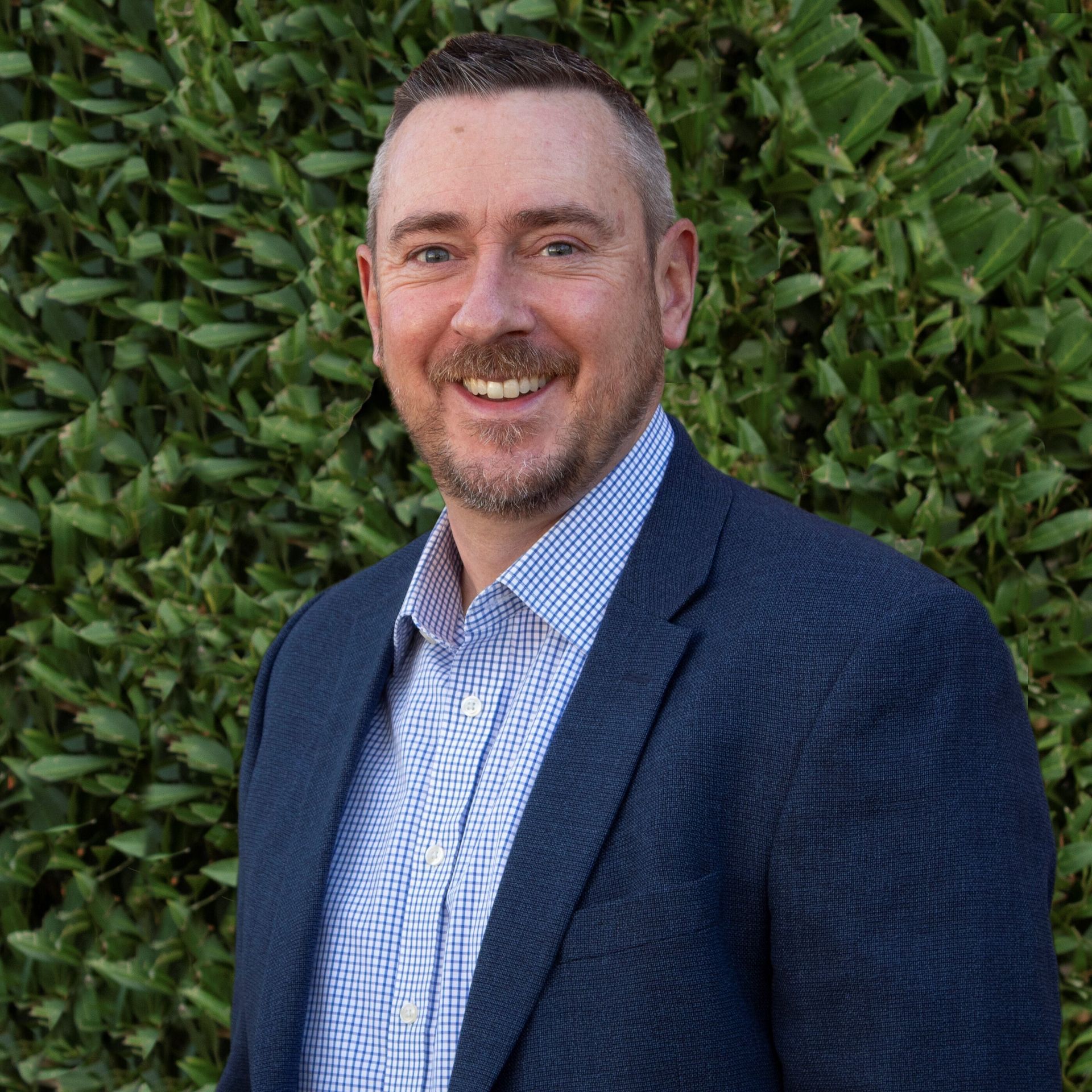 Man with a beard and graying hair, smiling, wearing a blue blazer and button-down shirt, in front of greenery.