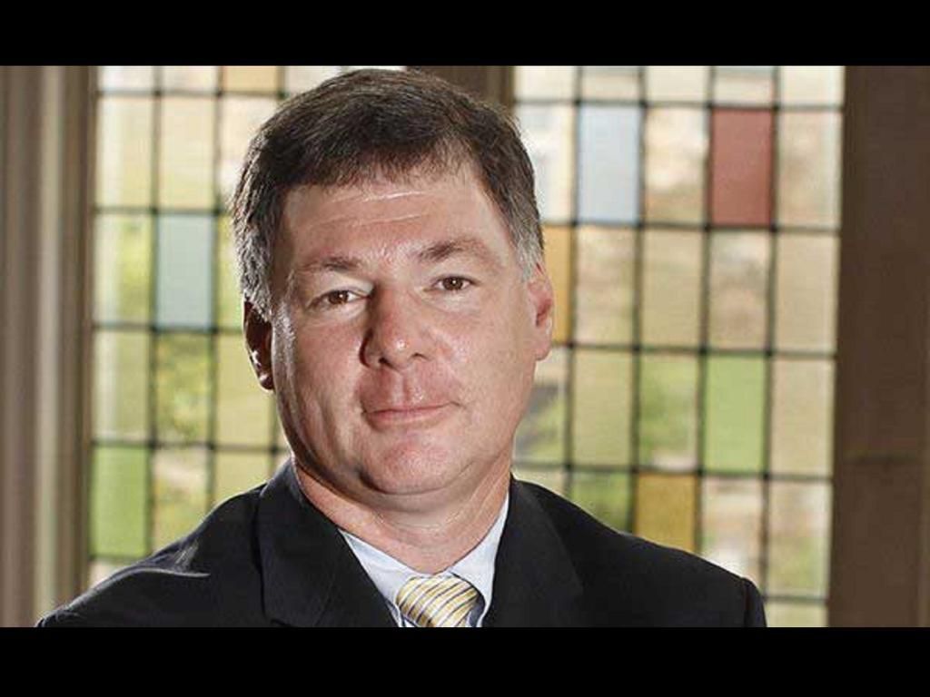 Man in suit and tie, smiling slightly, in front of a stained-glass window.