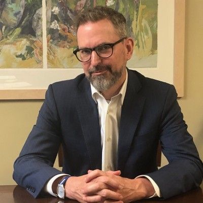 Man in glasses and navy suit, hands clasped, looking at camera. Painting in background.