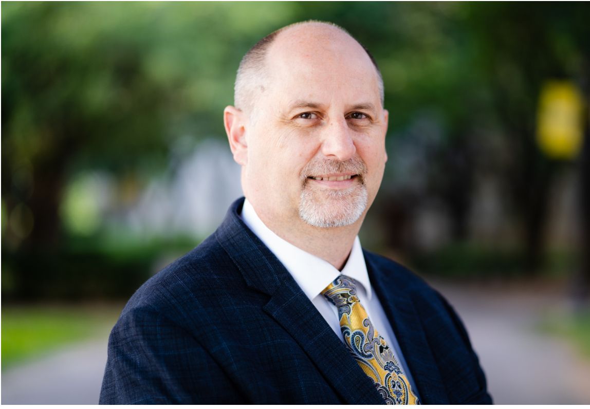 Man in suit and tie, smiling, outdoors on a path, trees in the background.