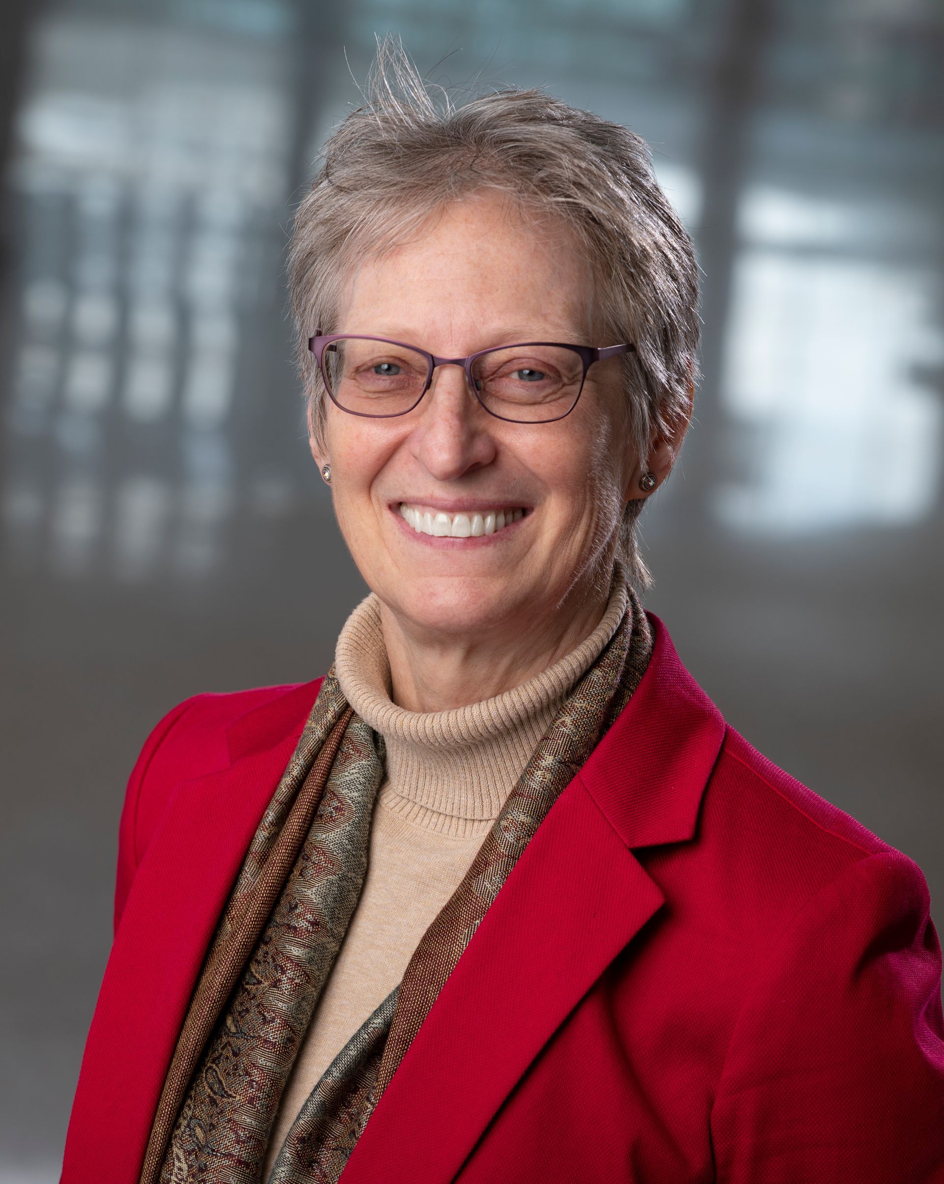 Woman with short gray hair, wearing glasses, a red jacket, smiling in a professional setting.