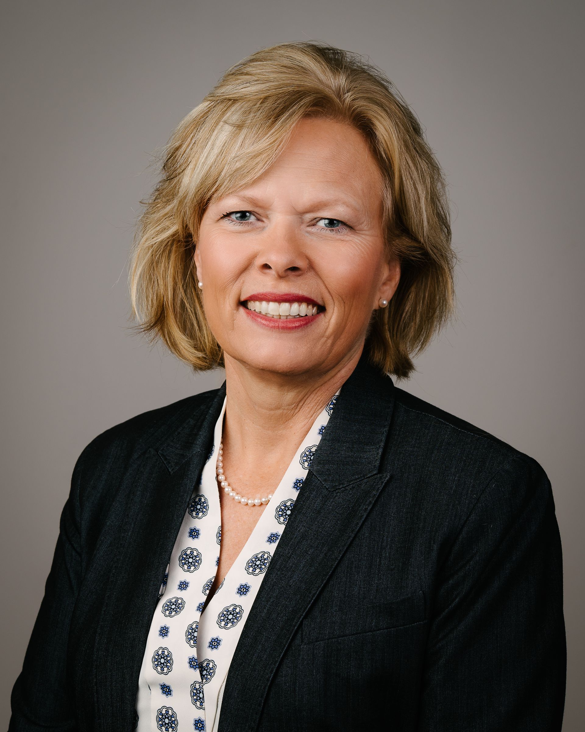 Woman in blazer smiles, posed headshot against a gray backdrop.