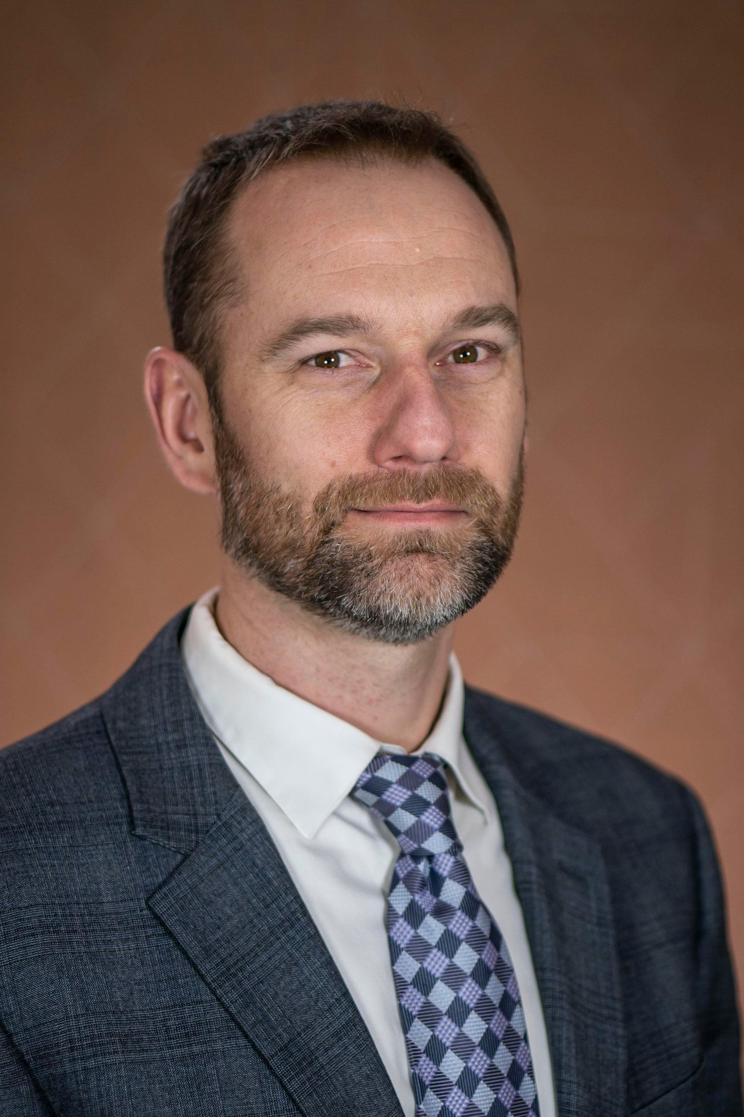 Man in suit and tie looking directly at the camera, neutral expression, against a brown background.