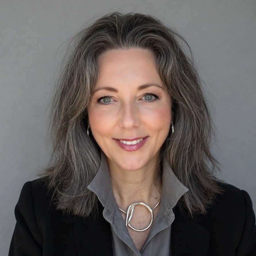 Woman with long gray hair, smiling, wearing a gray shirt, a black blazer, and silver necklace.