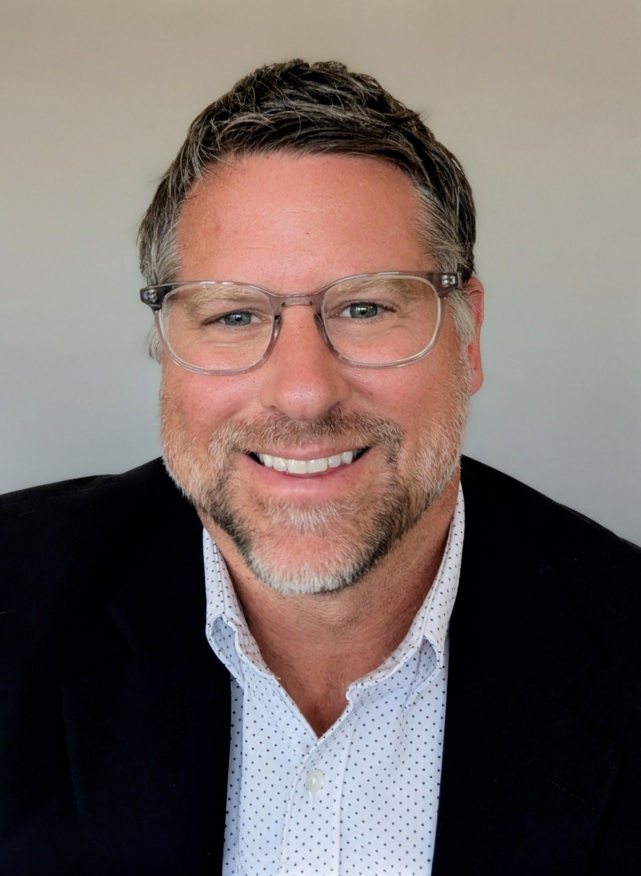 Man wearing glasses smiles, in a blazer over a button-down shirt.