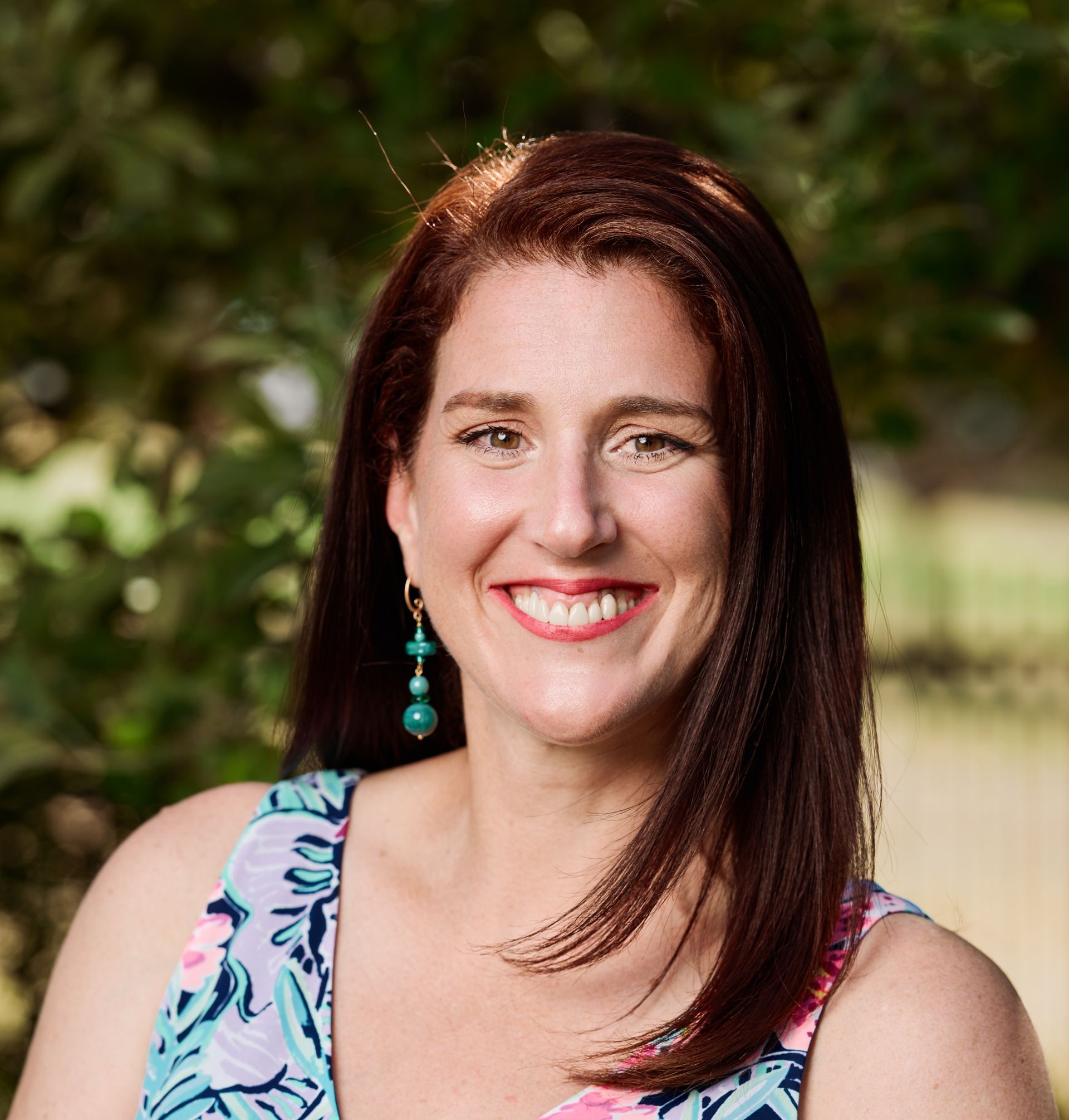 Woman with auburn hair smiling, wearing a patterned top and turquoise earrings, outdoors.