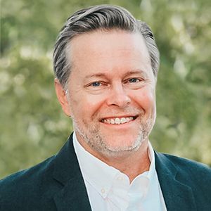 Man with short gray hair, smiling, wearing a dark blazer and white shirt. Outdoors with green foliage.