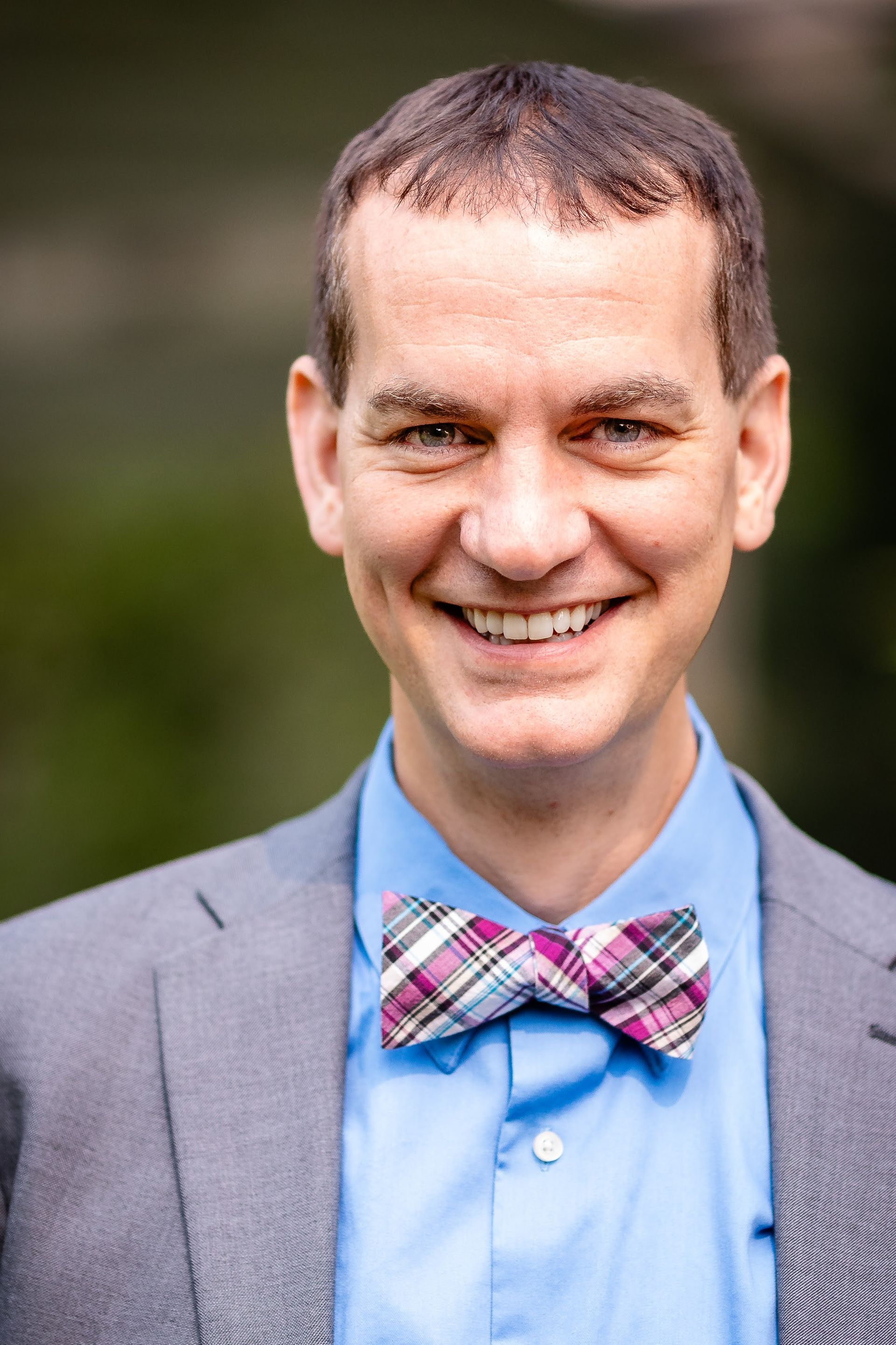 Man wearing a blue shirt, gray suit jacket, and plaid bow tie, smiling broadly outdoors.