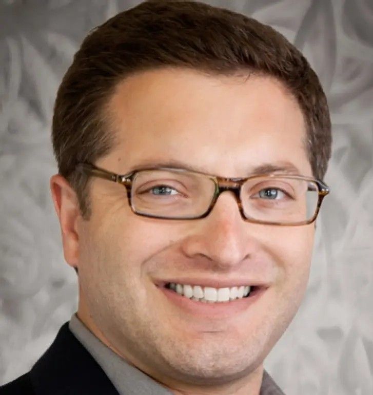Man with glasses smiles, wearing a dark blazer.