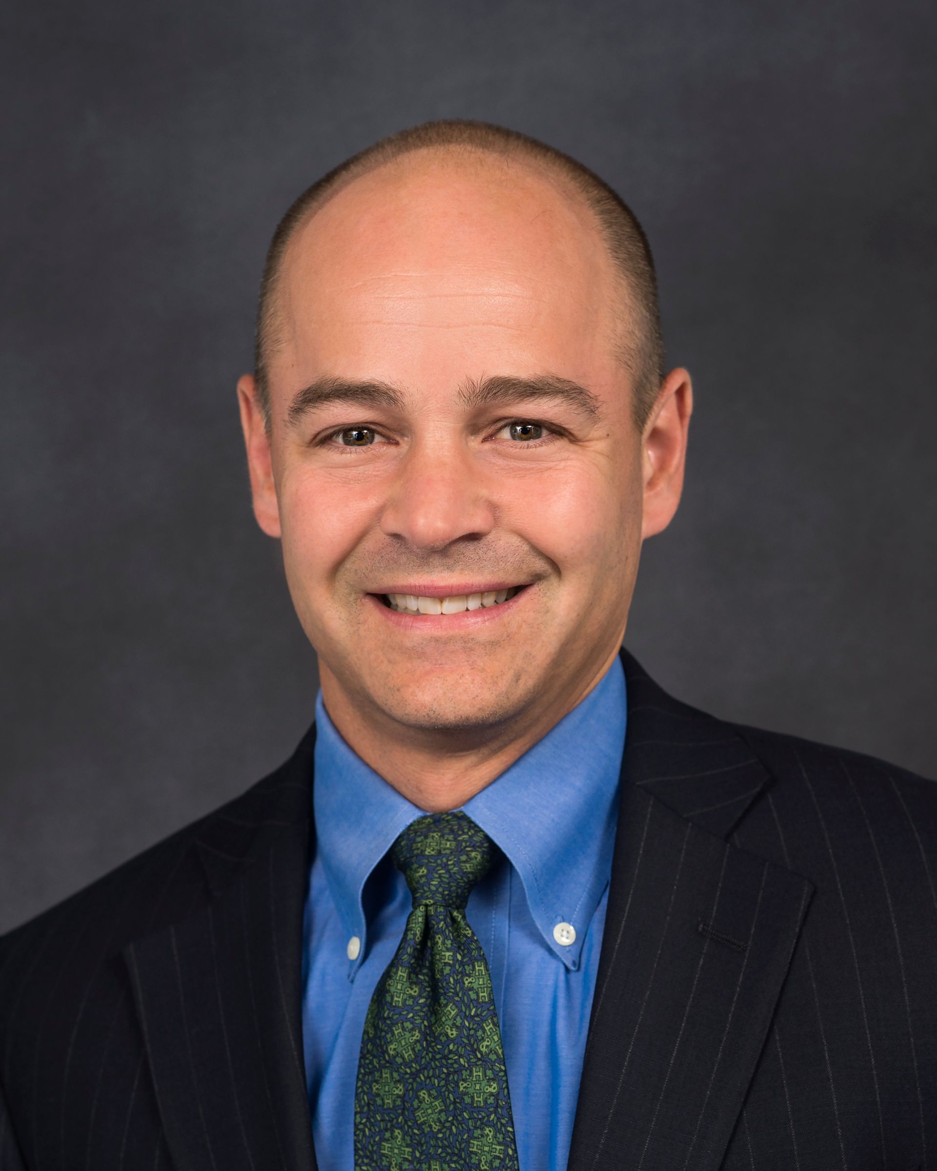 Man in blue shirt, green tie, and suit jacket smiles at the camera against a gray backdrop.