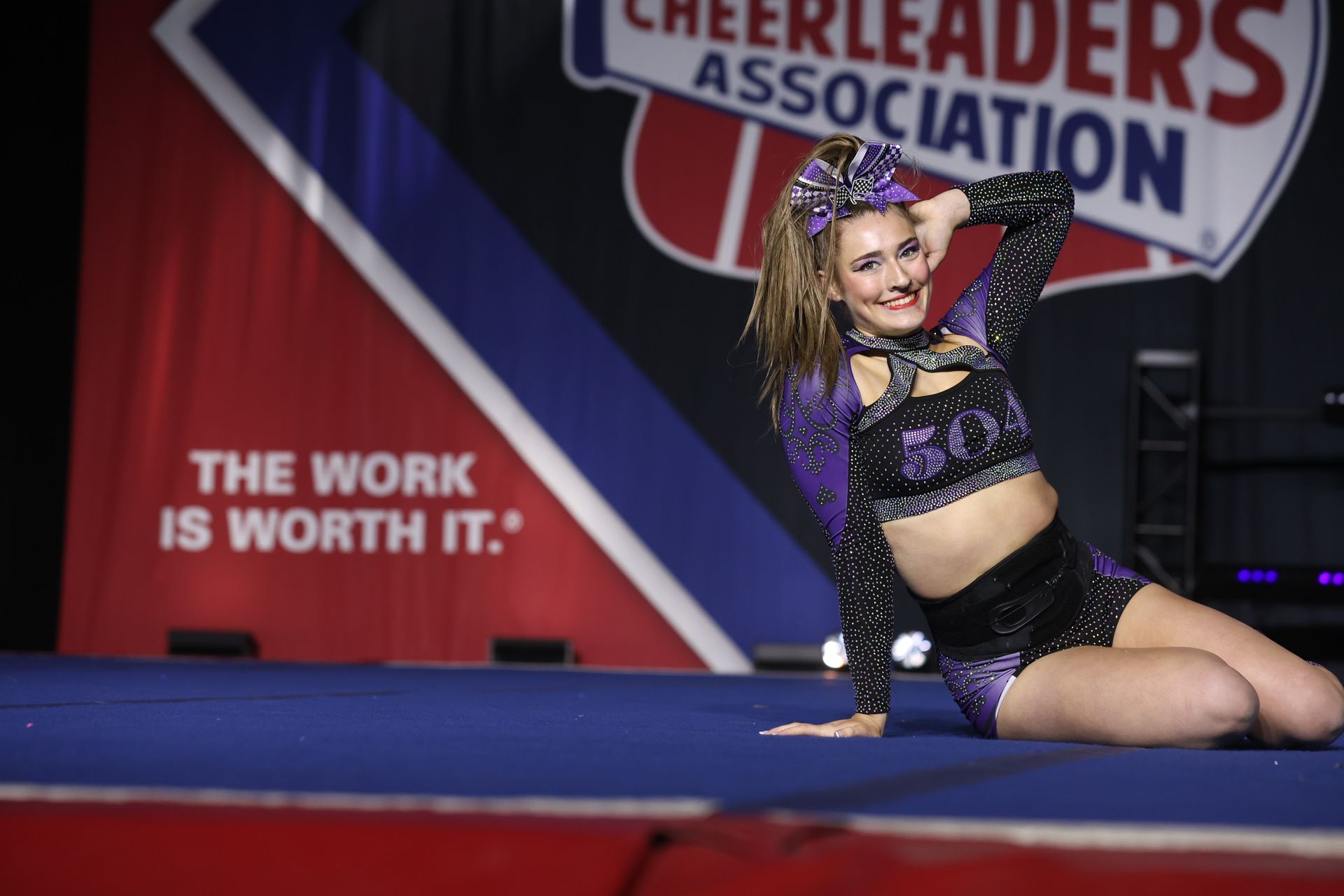 A cheerleader in a purple and black uniform posing on a blue competition mat in front of a banner.