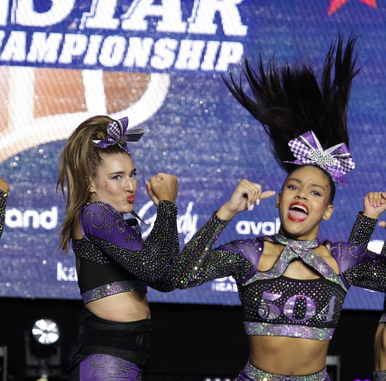 Two cheerleaders in purple and black uniforms with sparkling bows perform on stage in front of a championship banner.
