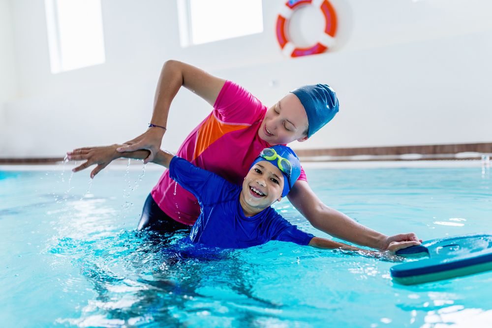 A swim instructor in a pink shirt guides a child in a blue shirt with a kickboard in a pool.