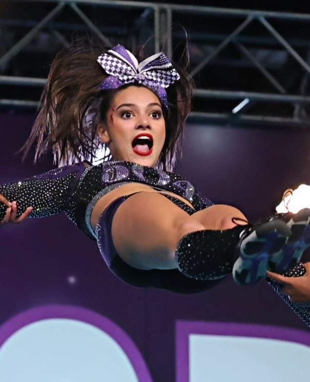 A cheerleader in a sparkling purple uniform is mid-air during a stunt, wearing a matching bow and looking surprised.