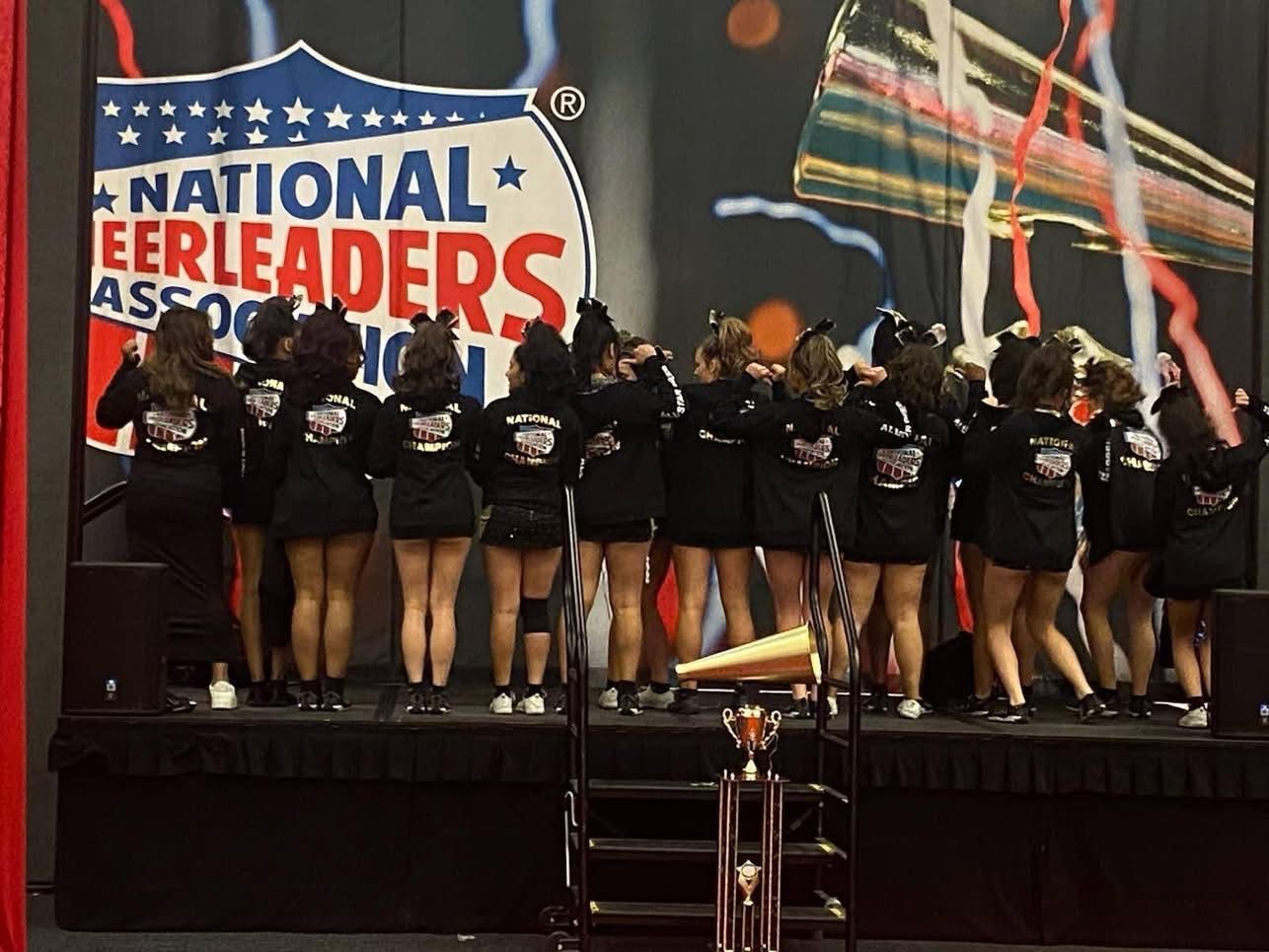 A cheerleading team stands on a stage in front of a National Cheerleaders Association banner with a trophy in front.