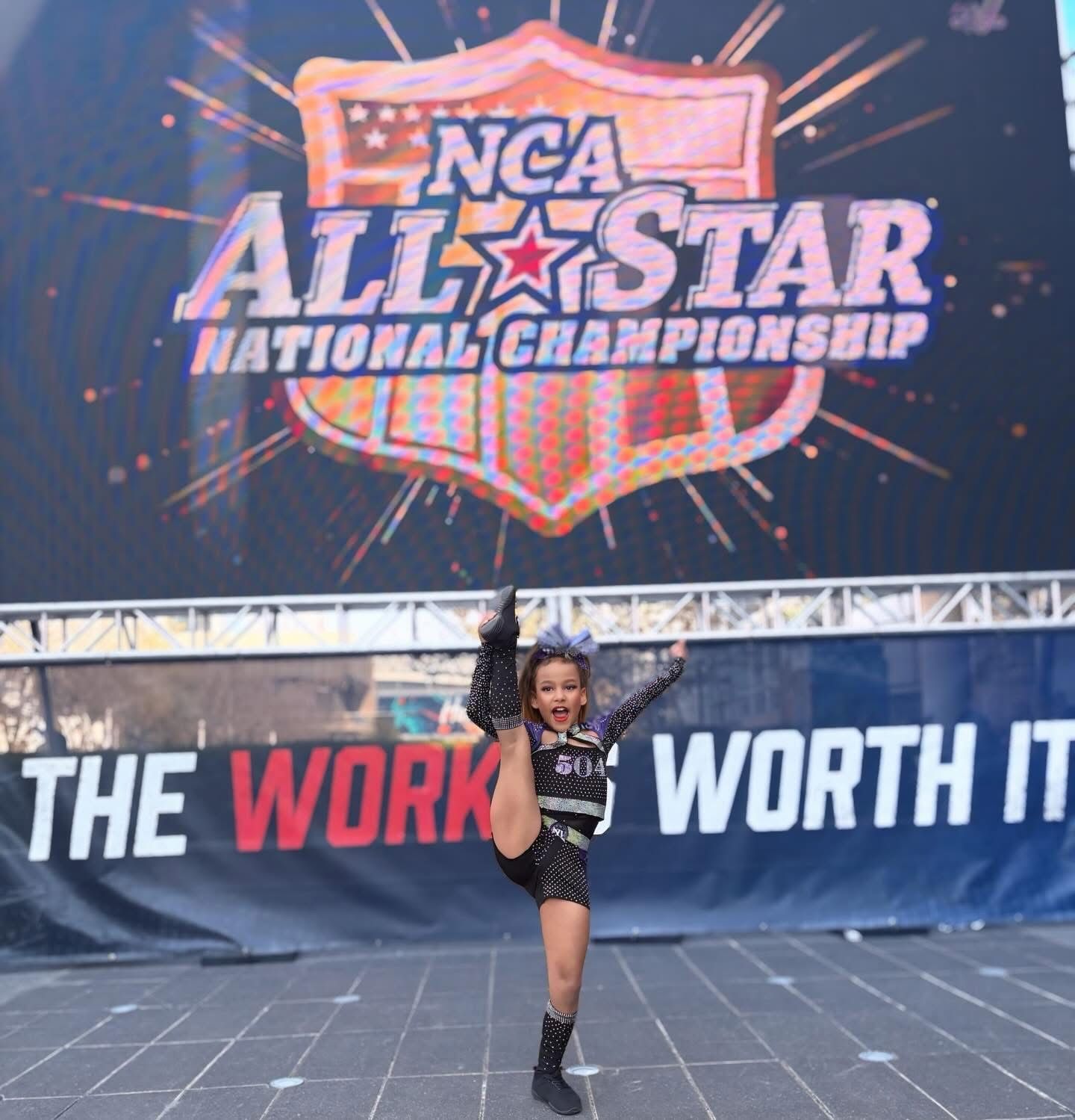 A cheerleader performing a scorpion stretch in front of an NCA All-Star National Championship banner.