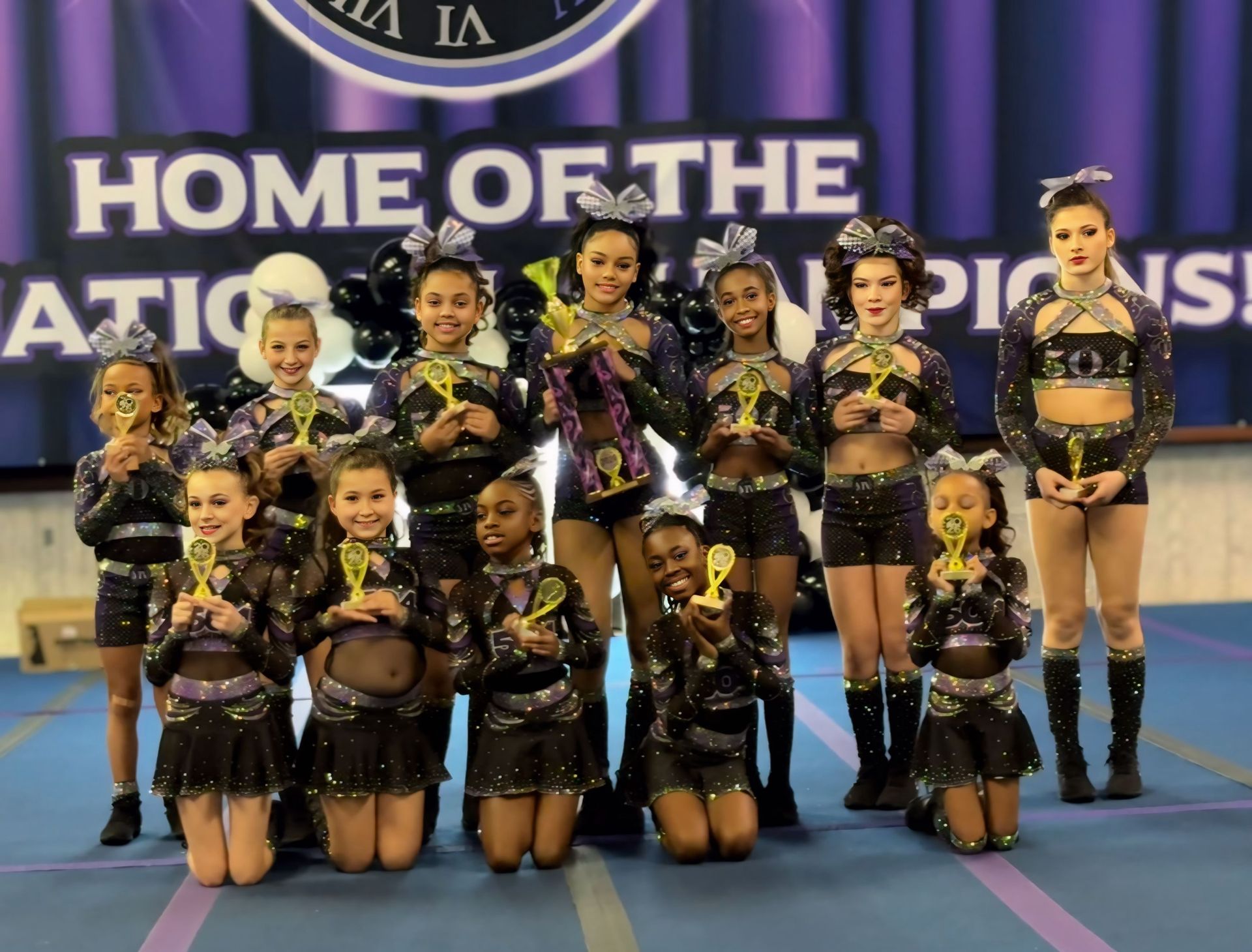 A group of cheerleaders in matching black uniforms holding gold trophies against a purple background.