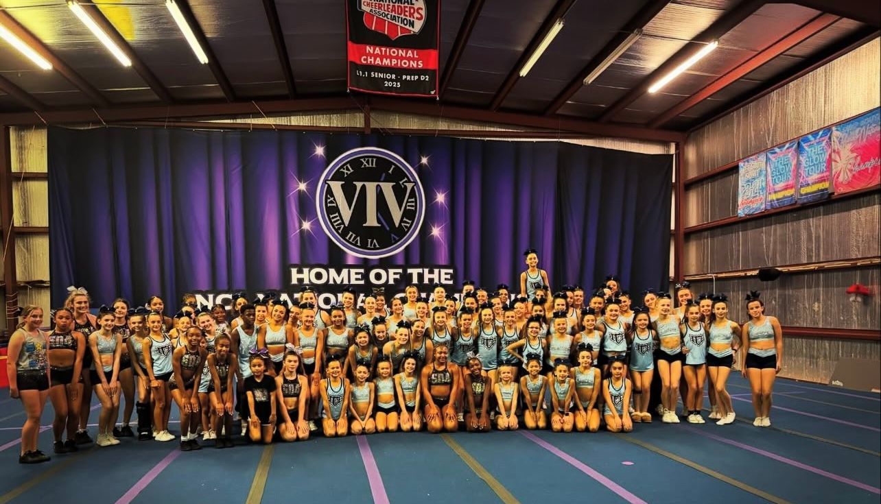 A cheerleading team in black uniforms stands on a stage in front of a National Cheerleaders Association banner.