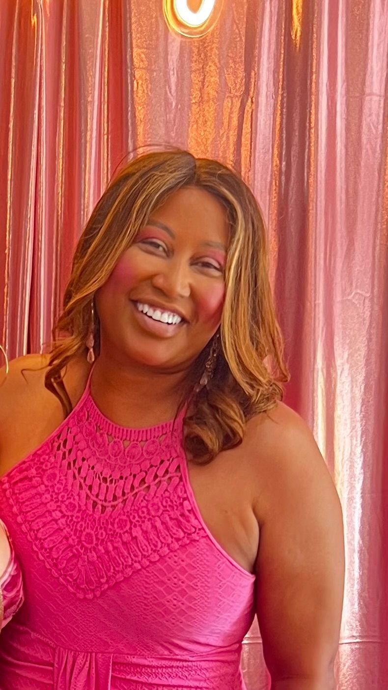 A smiling person with shoulder-length, highlighted hair wears a bright pink sleeveless top in front of a pink backdrop.