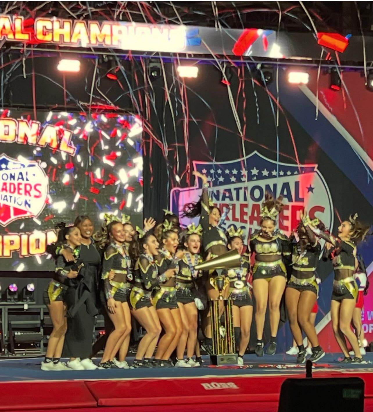 A cheerleading squad celebrates on a stage under falling confetti, holding a golden trophy as national champions.