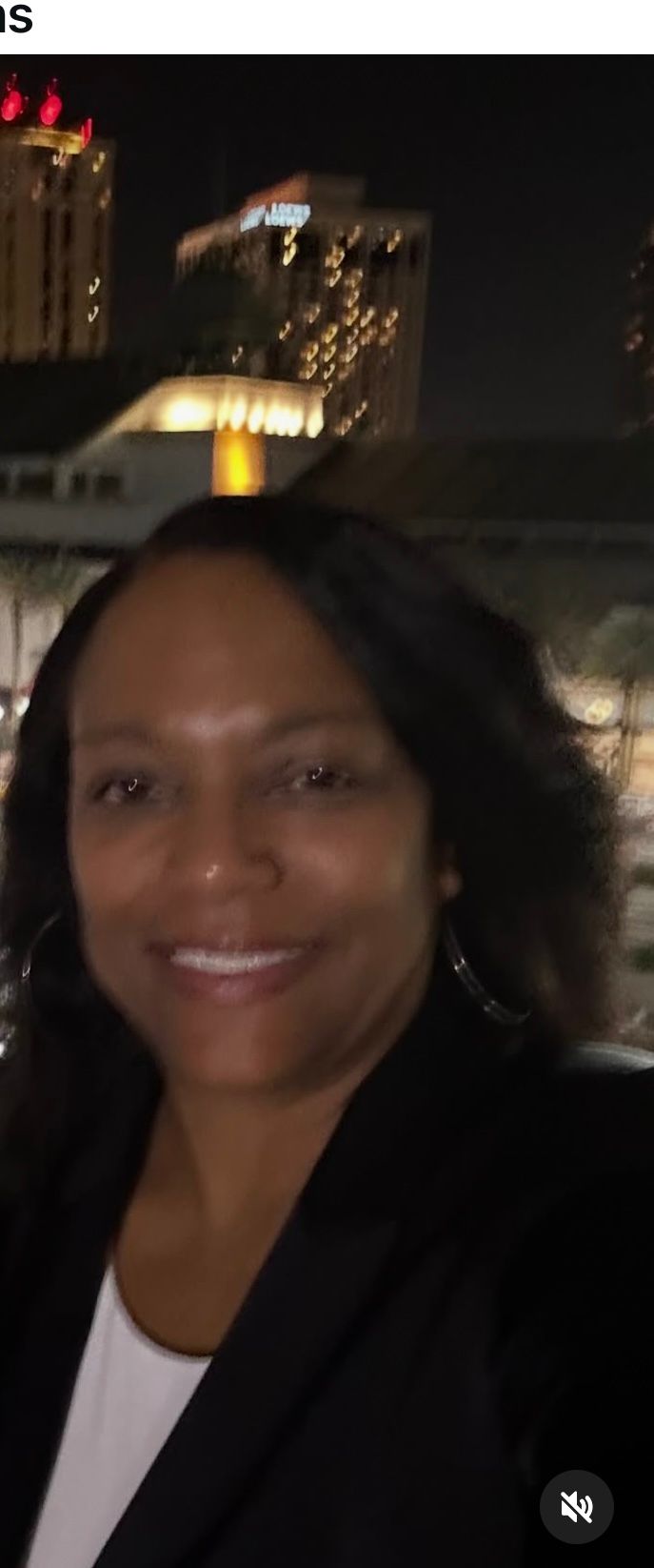 A person with dark hair smiling at the camera, wearing a dark blazer over a white top with a city skyline at night behind.