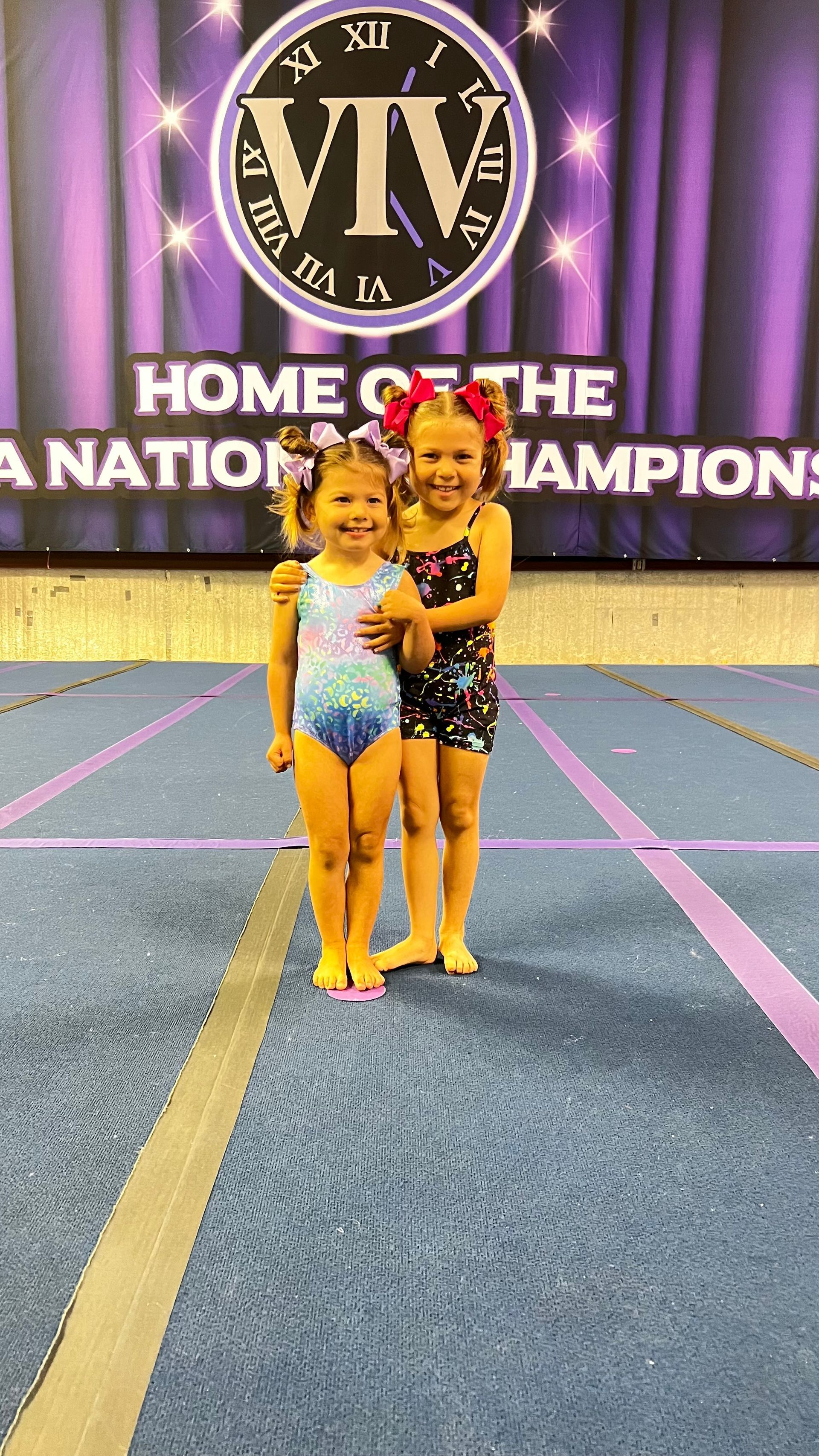 Two cheerleaders in purple and black uniforms with sparkling bows perform on stage in front of a championship banner.